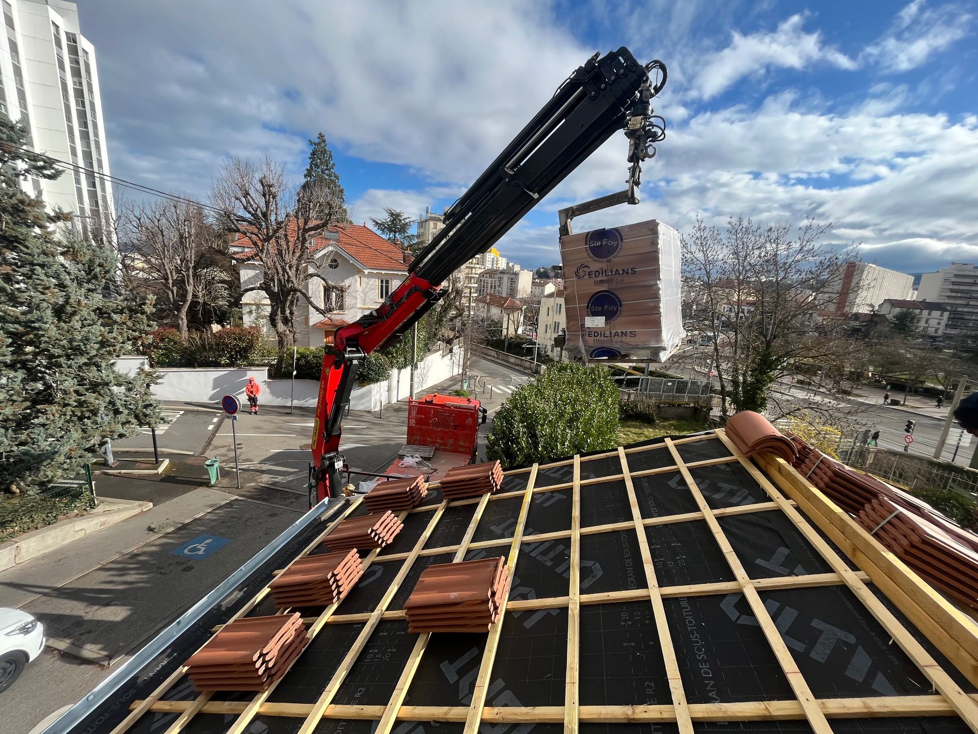 Une grue dépose des matériaux de toiture sur un bâtiment en construction, avec des ouvriers et des arbres en arrière-plan.