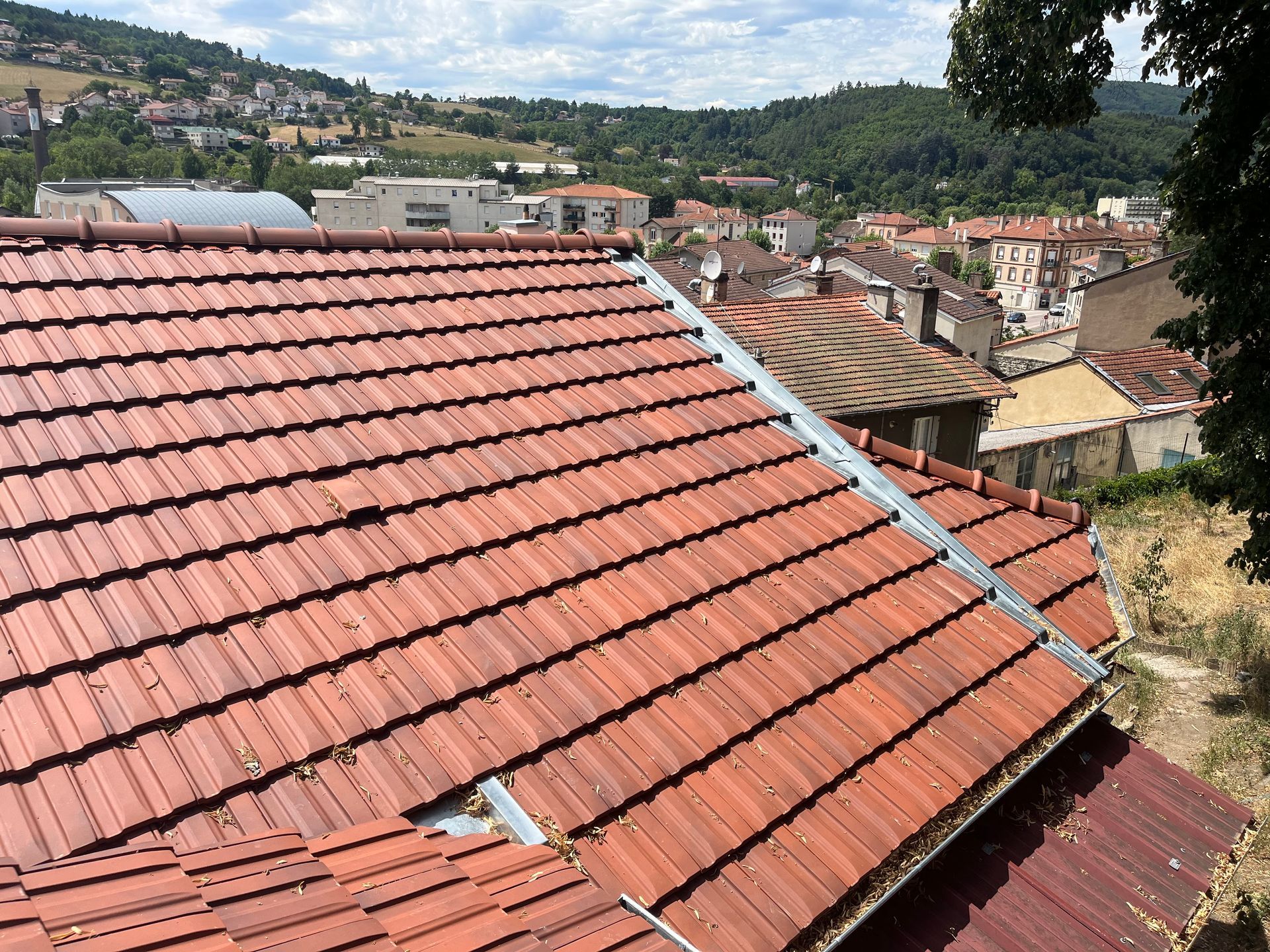 Un toit de tuiles rouges surplombe une ville aux immeubles de plusieurs étages et aux collines lointaines, sous un ciel partiellement nuageux.