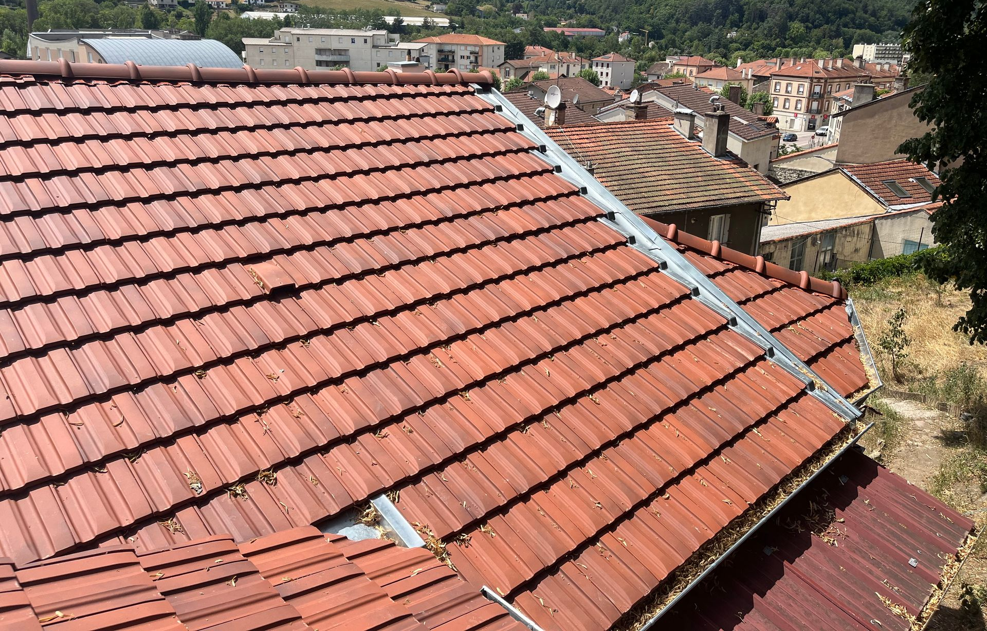 Toit en pente de tuiles rouges donnant sur une ville à flanc de colline, avec des bâtiments et de la verdure en arrière-plan.