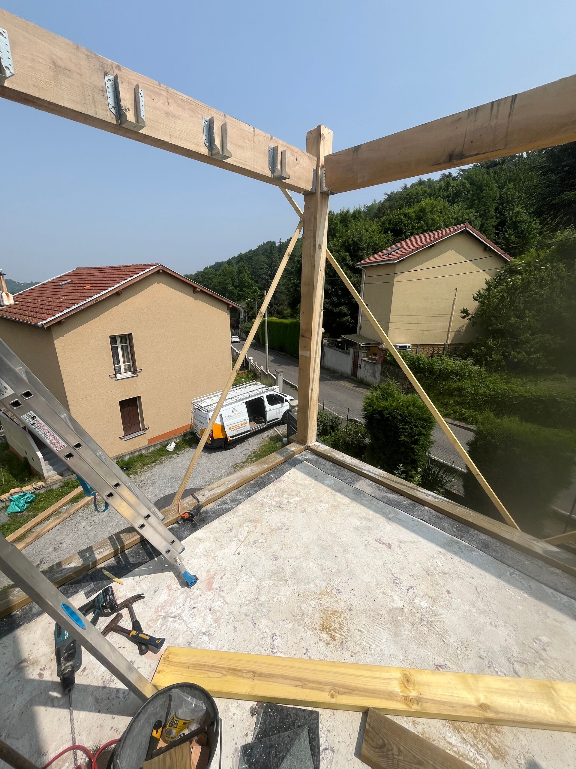 Vue d'un chantier de construction sur un toit, avec des poutres en bois, une échelle et des maisons en contrebas par une journée ensoleillée.