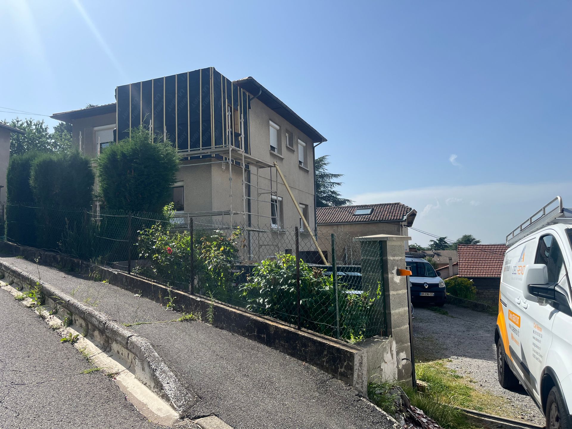 Maison en pierre avec allée en pente, verdure et voitures garées sous un ciel bleu azur.