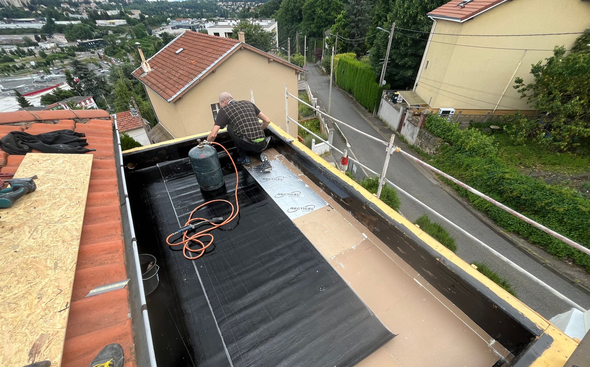 Toiture en construction avec toiture noire, outils et deux ouvriers sur un toit près de maisons et d'une route.