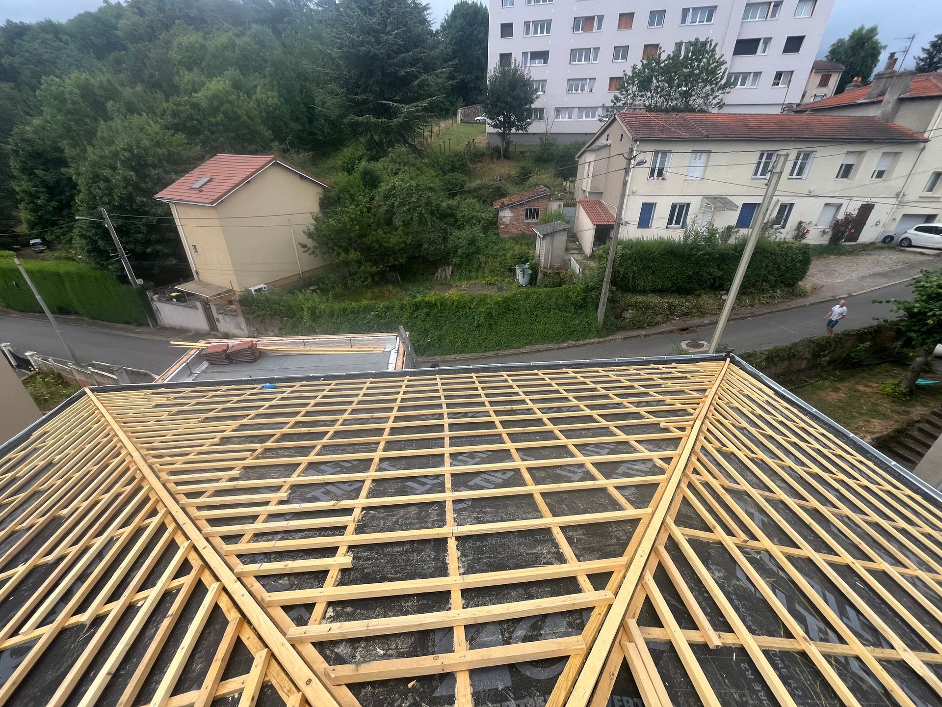 Charpente de toit en construction donnant sur des maisons et une route dans un quartier résidentiel boisé.