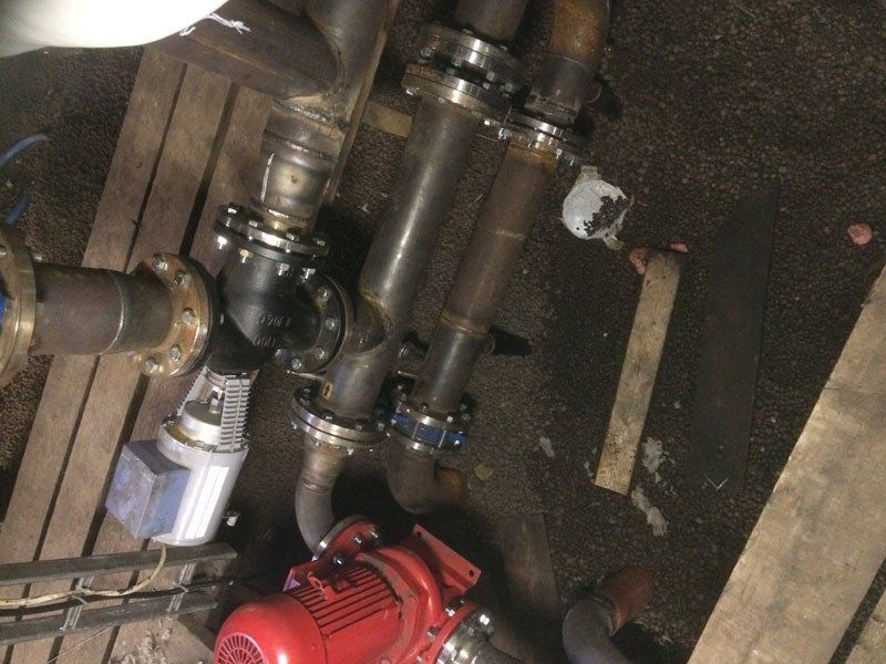 Punainen sähköpumppu kytkettynä sarjaan teollisuuskäyttöön tarkoitettuja metalliputkia ja laippoja hämärässä, pölyisessä kodinhoitotilassa.