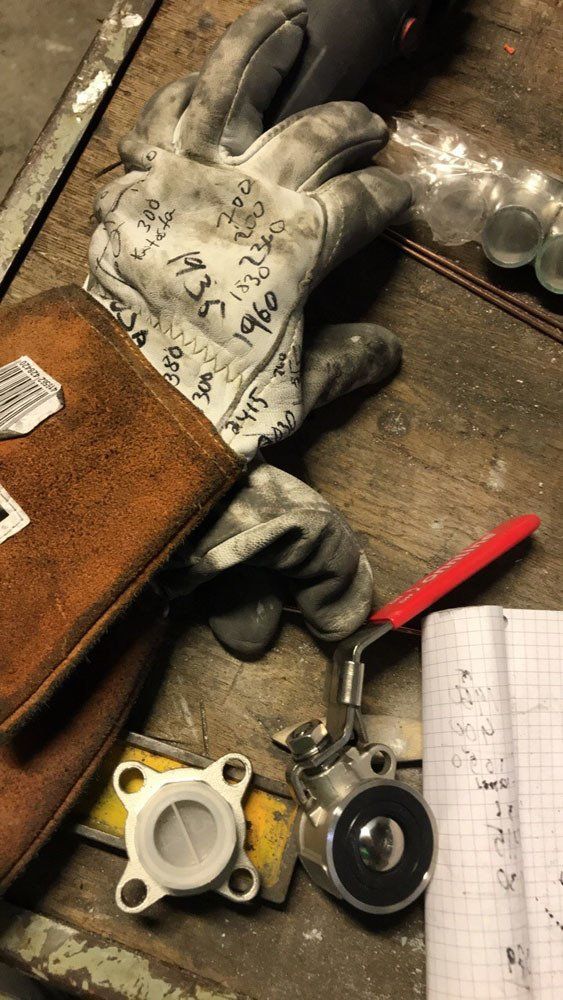 Työpöytä, jossa on hitsauskäsine, pieni nelireikäinen metalliliitin, kuulaventtiili ja ruutupaperivihko.