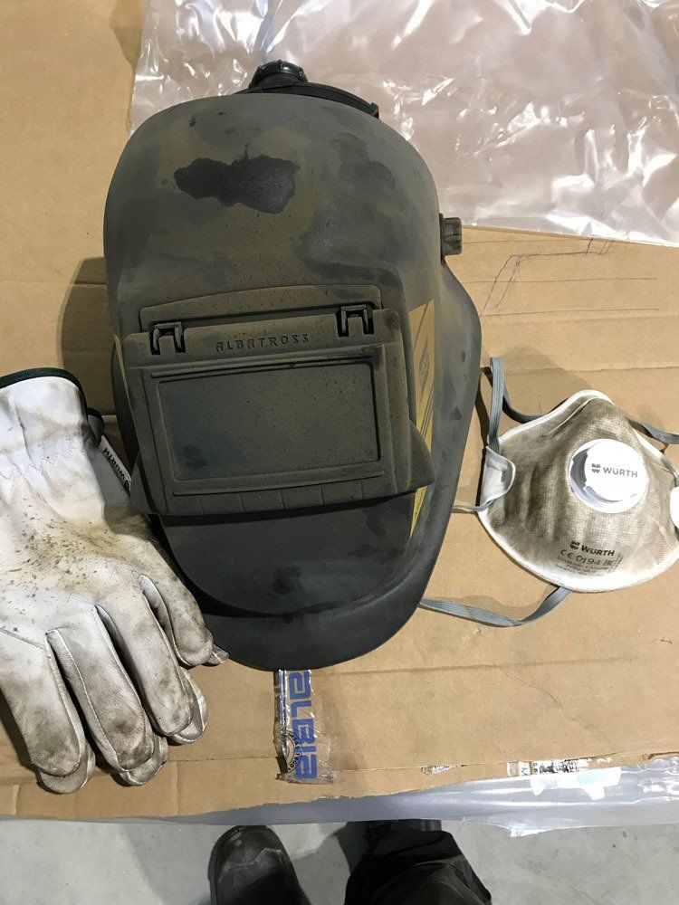 Hitsauskypärä, pari nahkaisia ​​työkäsineitä ja kertakäyttöinen pölynaamari pahvipinnalla.