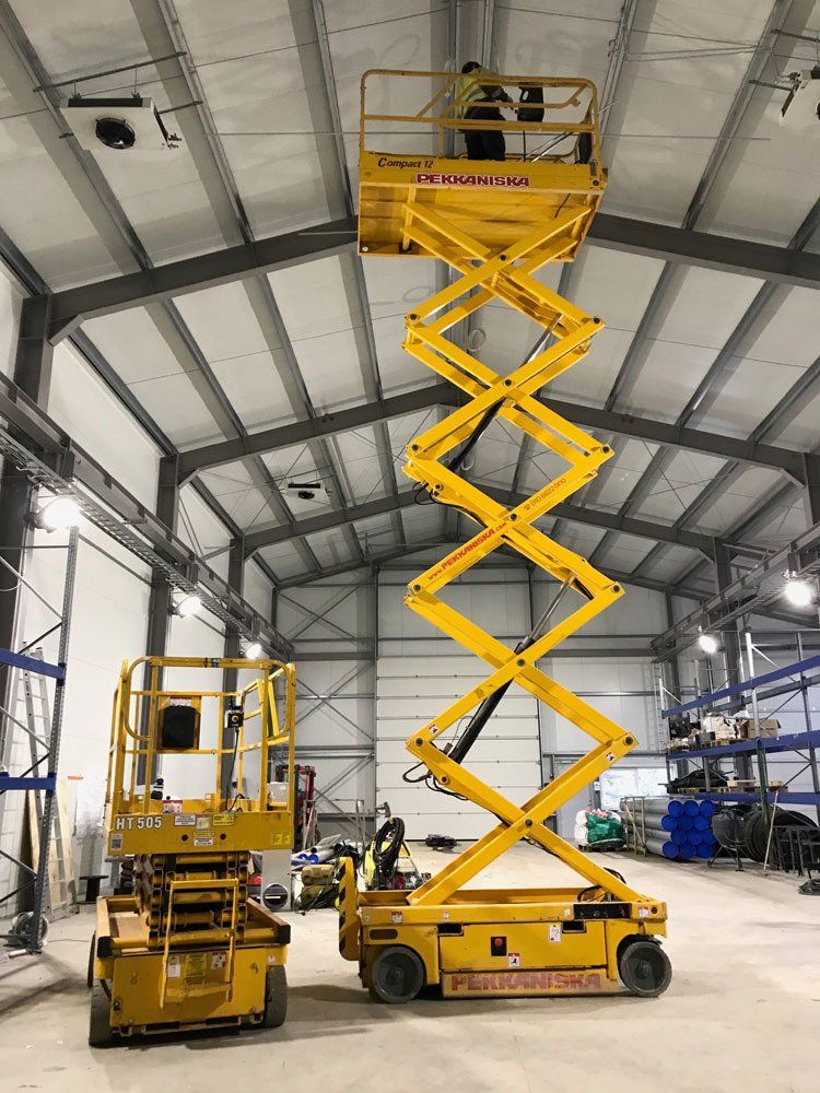 Henkilö nostetulla keltaisella saksinostimella varastossa, ja lähellä on toinen, sisäänvedetty saksinostin.