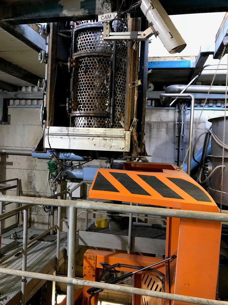 Teollisuuskoneita, joissa on suuri metalliverkkosylinteri ripustettuna kirkkaan oranssin turvasuojuksen yläpuolelle varastossa.