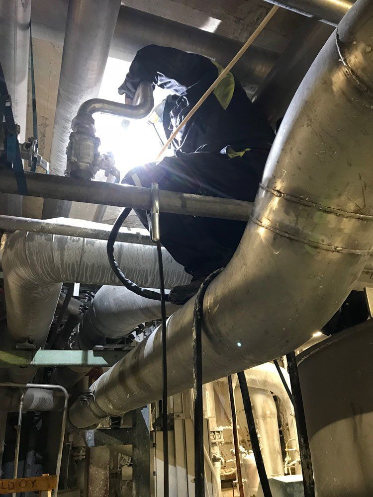 Suojavaatetukseen pukeutunut työntekijä hitsaa putkea korkealla teollisuustilassa, joka on täynnä metallisia koneita.