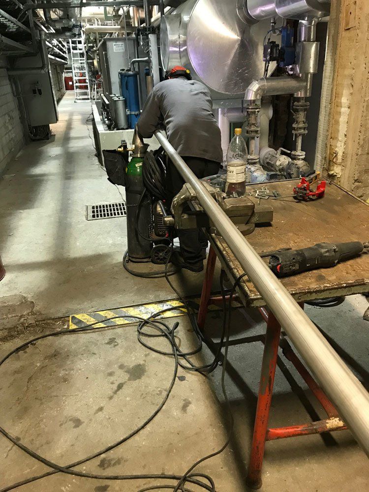 Harmaapaitainen työntekijä istuu työpöydän ääressä kodinhoitohuoneessa ja hitsaa pitkää metalliputkea, joka on kiinnitetty ruuvipenkkiin.