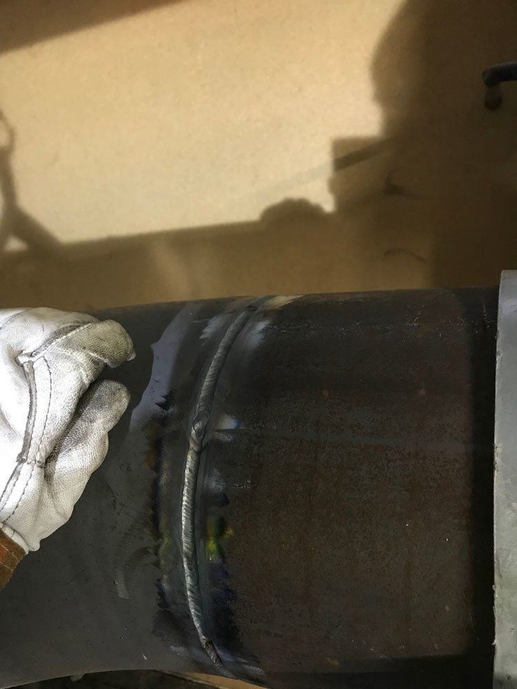 Lähikuva hansikaspäisestä kädestä vastavalmistuneen metalliputkihitsauksen vieressä, jossa näkyy tumma, kuvioitu liitos.