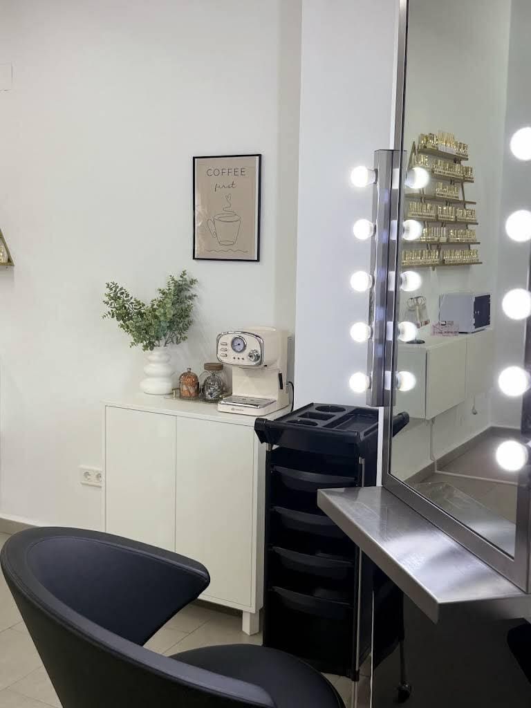 Estación de maquillaje de salón con espejos iluminados, silla negra, armario blanco y plantas en una habitación moderna.