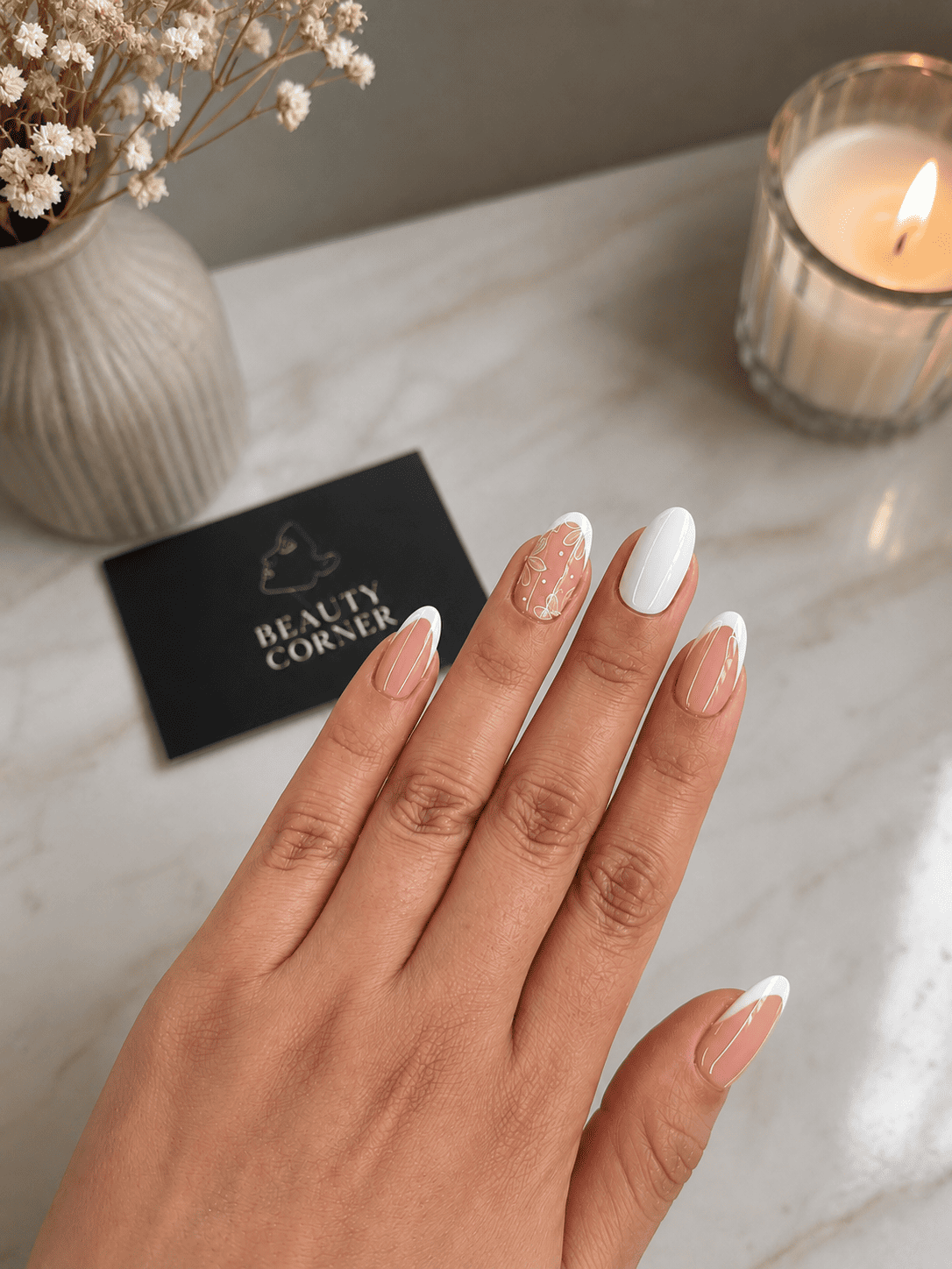 Mano bien cuidada con uñas blancas sobre una superficie de mármol junto a una vela y flores.