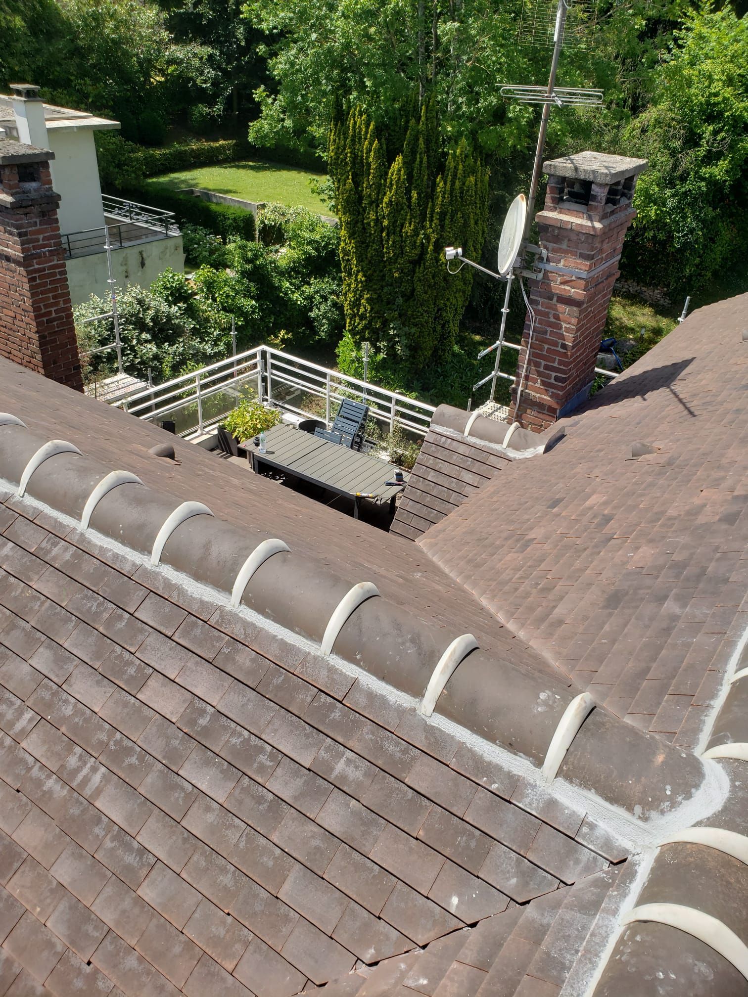 Vue d'un toit de tuiles avec cheminée, antenne et petit patio avec de la verdure en arrière-plan.