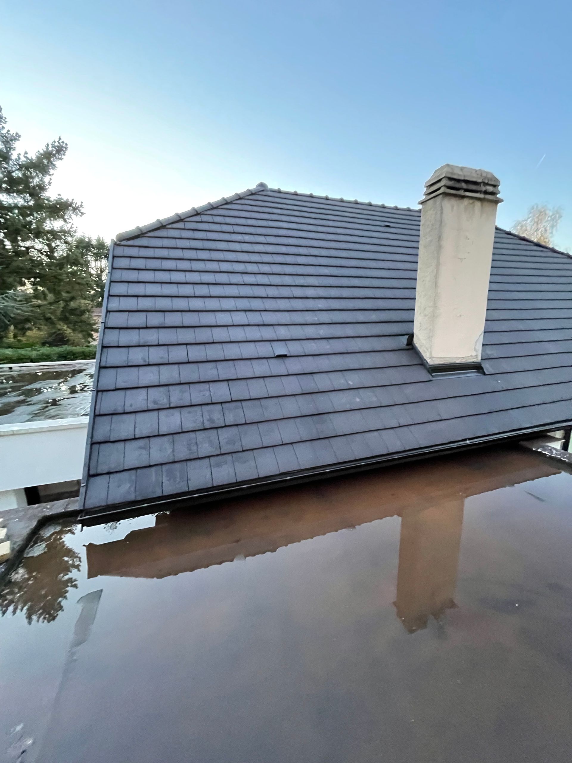 Un toit de bardeaux sombres et une cheminée blanche surplombent une surface recouverte d'eau, avec un ciel bleu clair en arrière-plan.