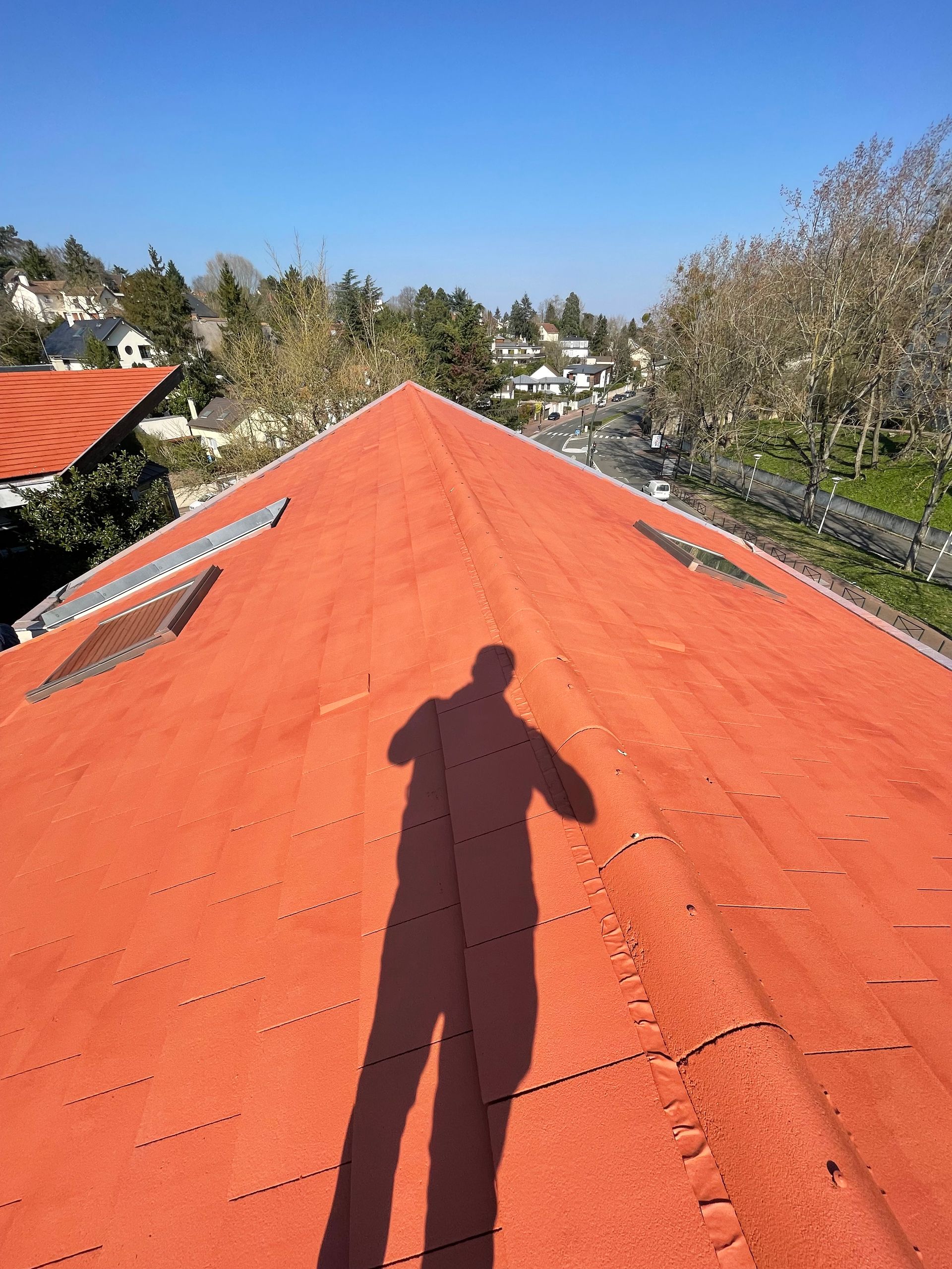 Ombre d'une personne sur un toit rouge sous un ciel bleu, avec des arbres et des bâtiments en arrière-plan.