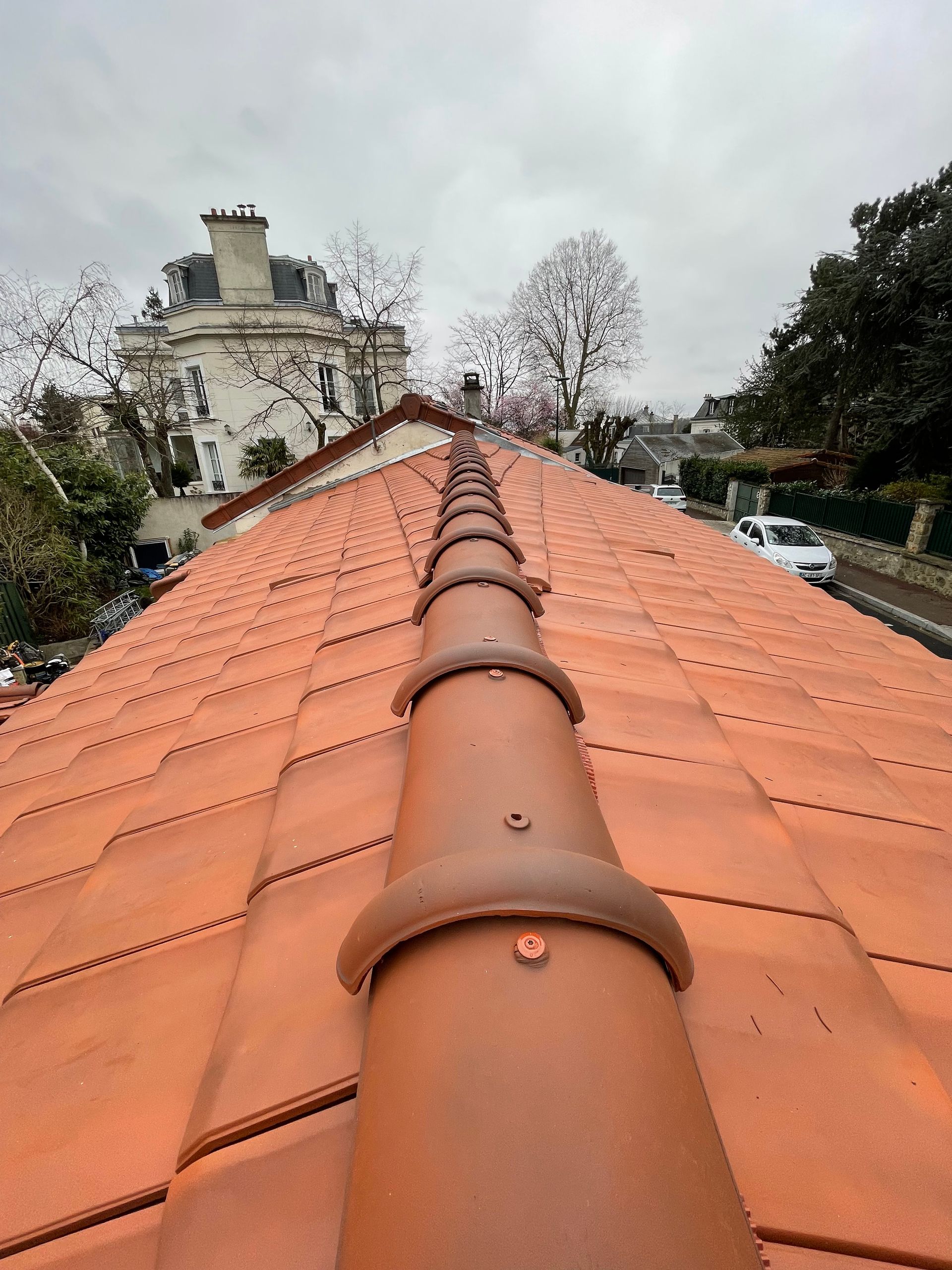 Vue depuis le faîte du toit ; tuiles de terre cuite rouges, tuiles faîtières brunes, ciel nuageux, bâtiments en arrière-plan.