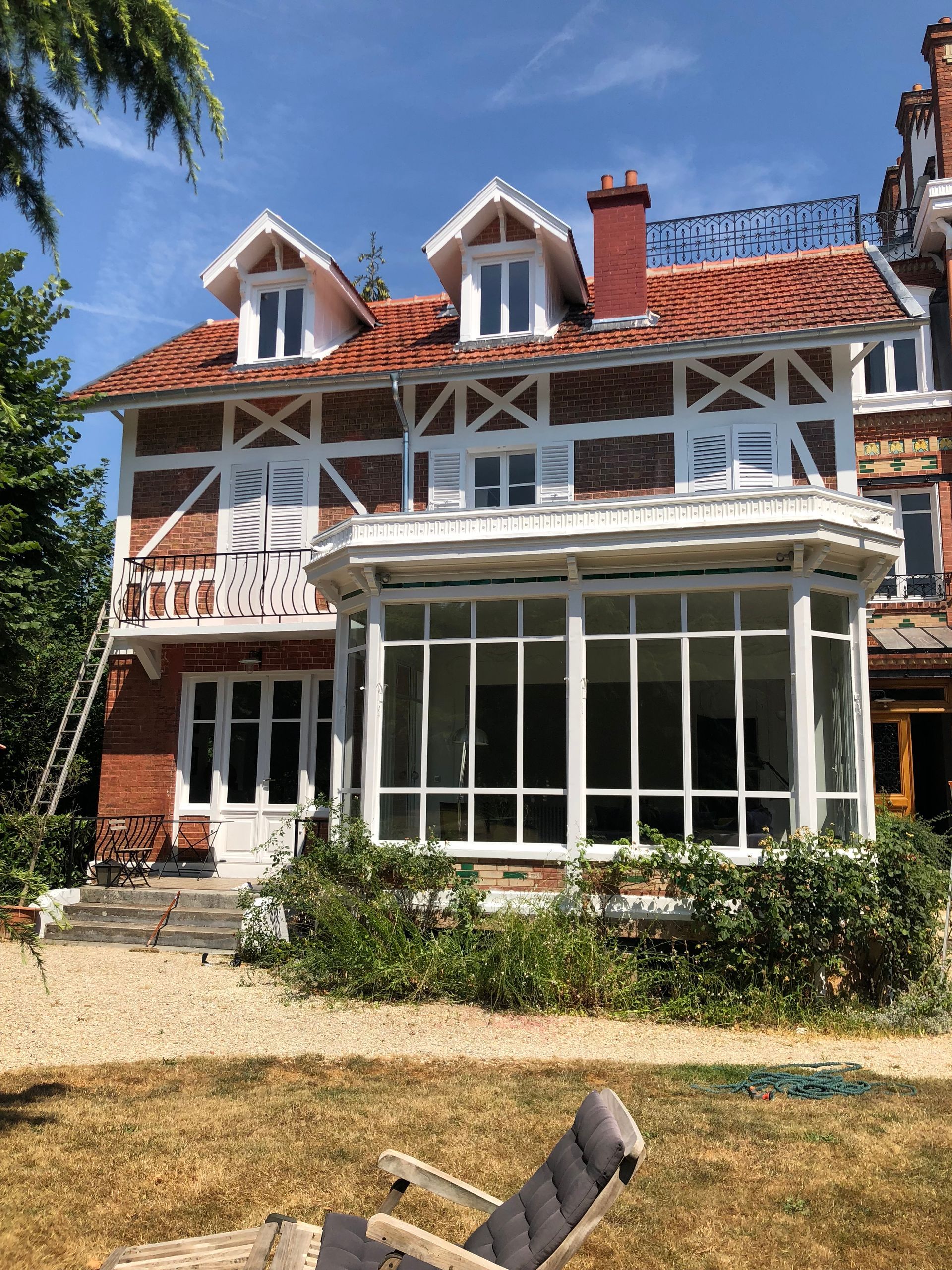 Maison en briques de deux étages avec des boiseries blanches, une baie vitrée et un toit de tuiles rouges, se détachant sur un ciel bleu.