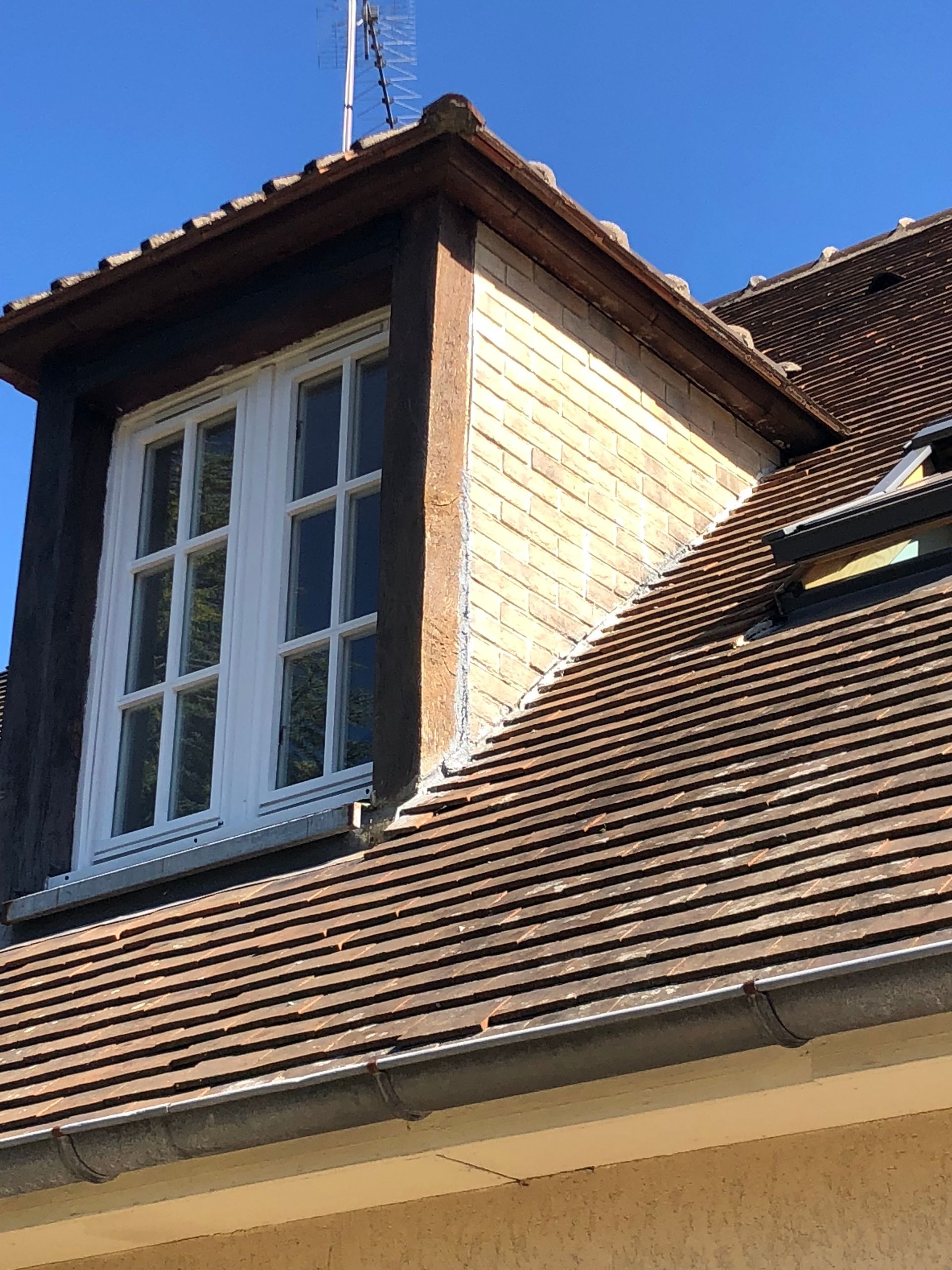 Lucarne à cadre blanc, sur un toit de tuiles. Boiseries brunes. Ciel bleu.