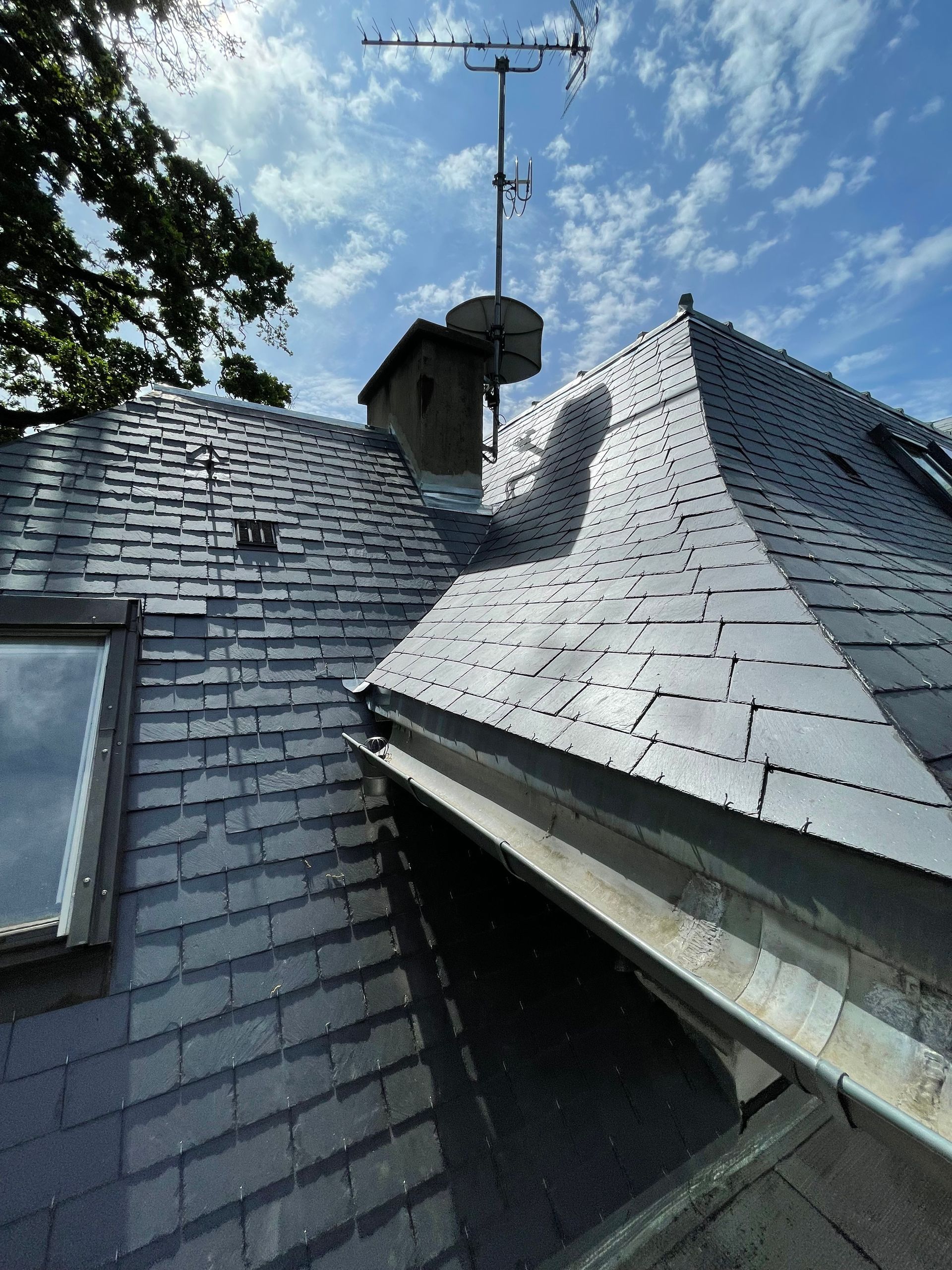 Toit en ardoise avec lucarne, cheminée et antenne de télévision sur fond de ciel bleu.