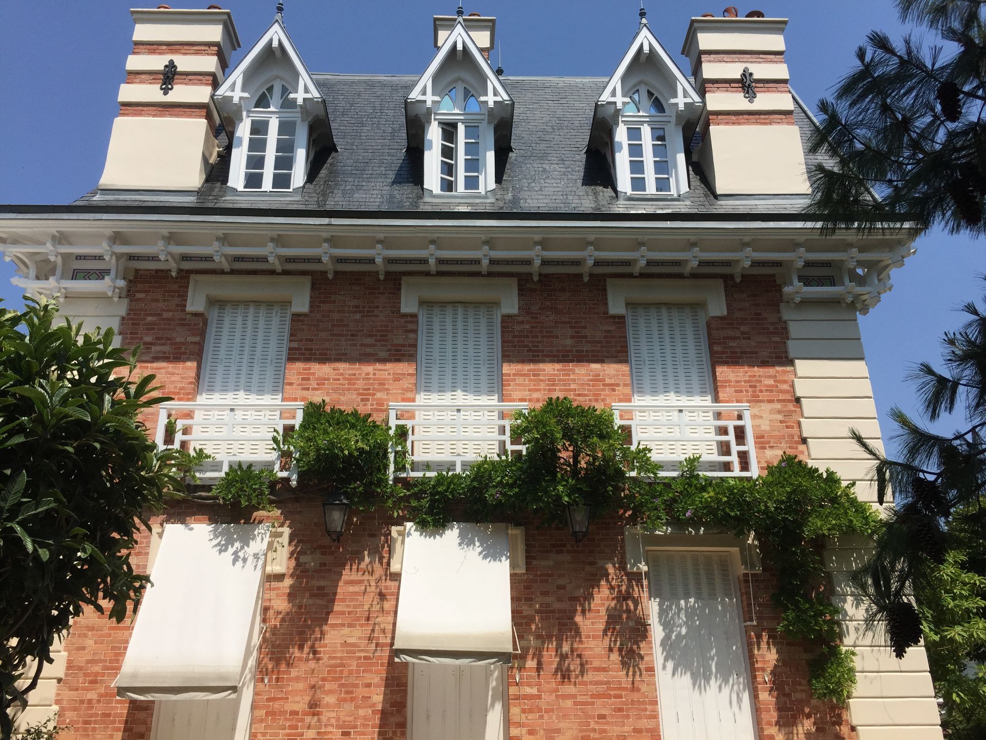 Maison en briques au toit sombre et aux boiseries blanches. Trois lucarnes. Balcon recouvert de vigne. Ciel bleu.