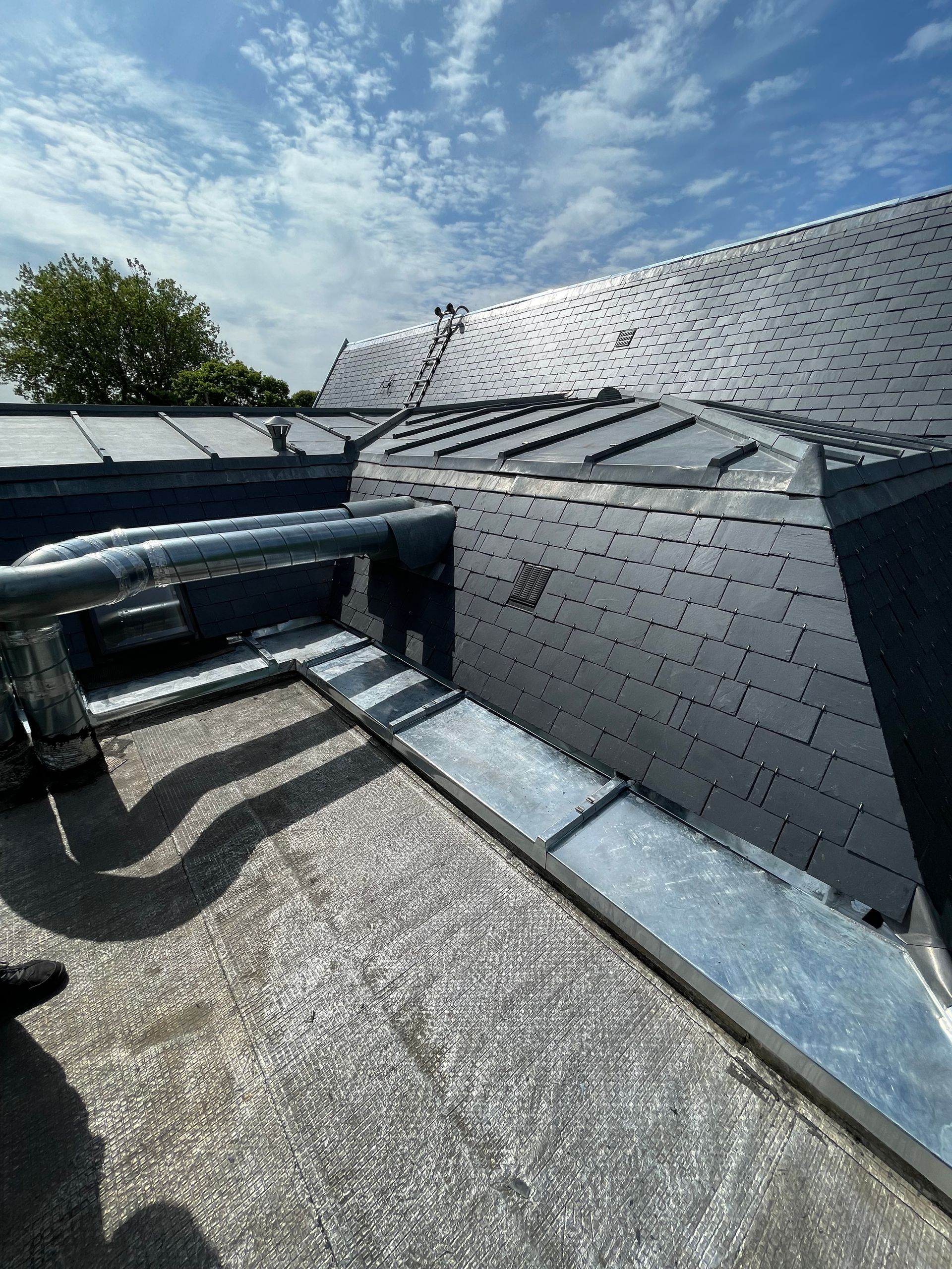 Vue d'un toit de tuiles sombres avec des solins métalliques et des tuyaux de ventilation par une journée ensoleillée.