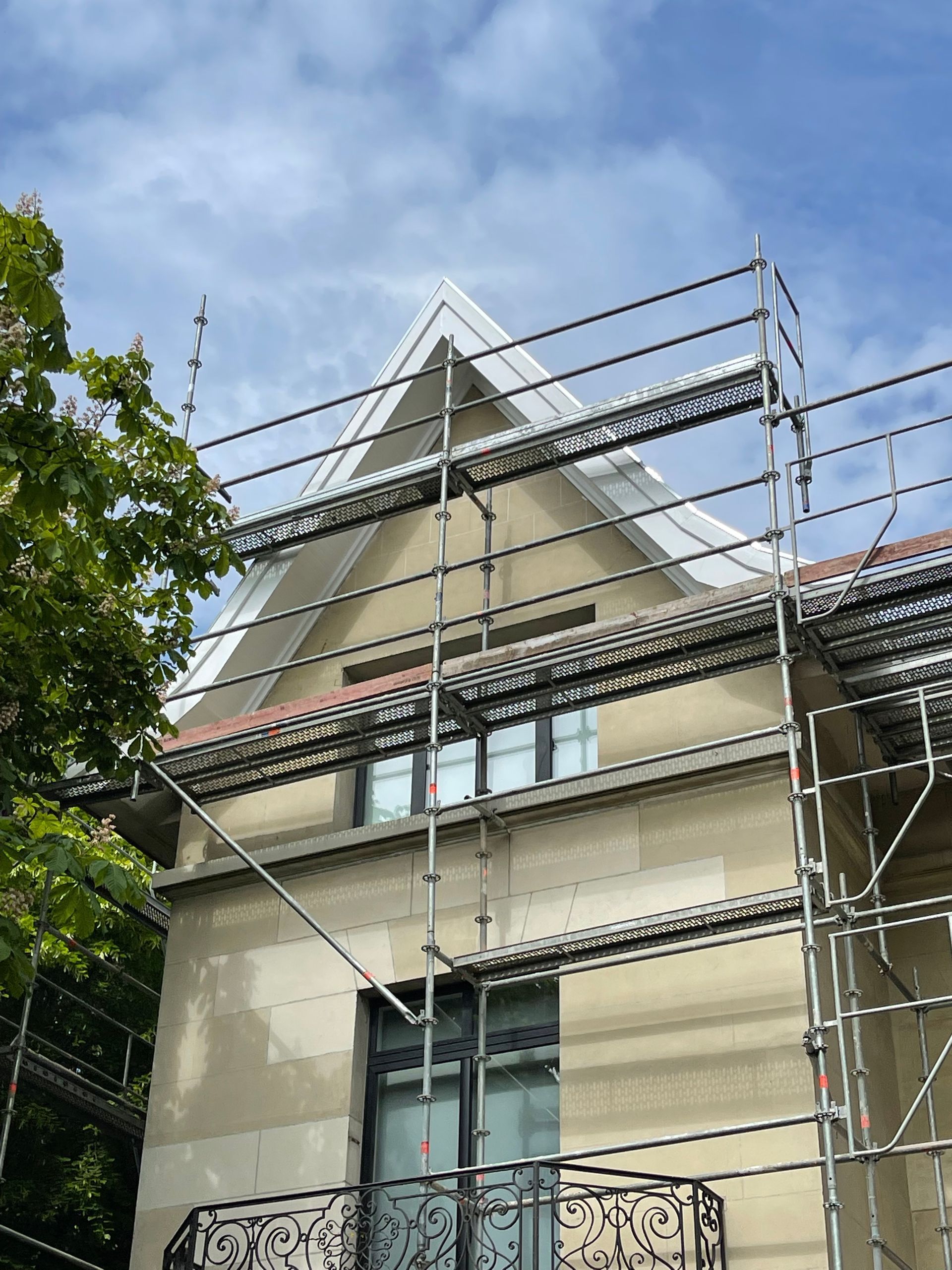Façade d'immeuble en réparation, échafaudages, toit triangulaire avec des accents gris et rouges, ciel bleu.