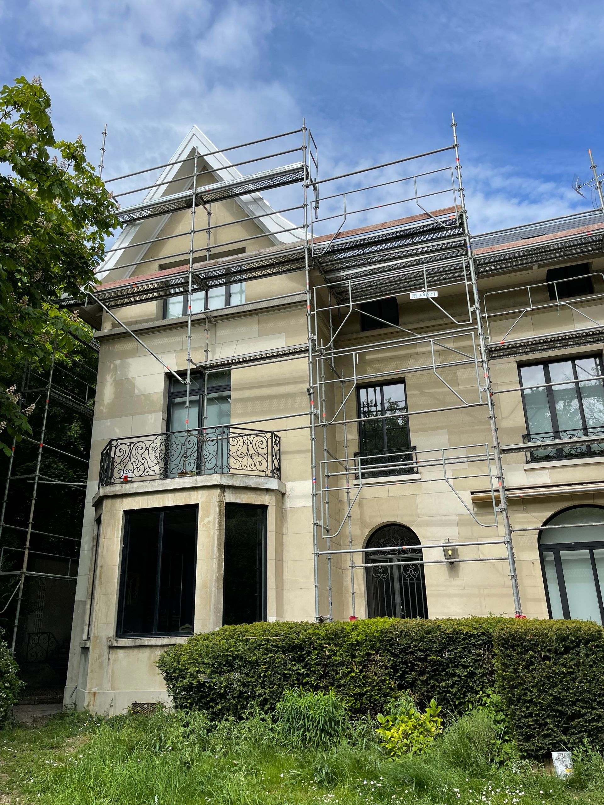 Des échafaudages entourent un immeuble de plusieurs étages à la façade claire ; la végétation y est envahissante au premier plan.
