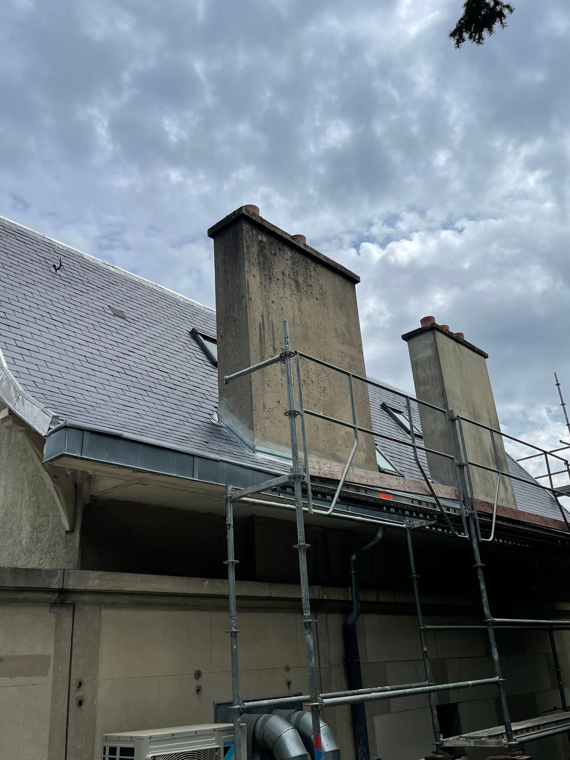 Toit avec deux cheminées, échafaudages et ciel nuageux.