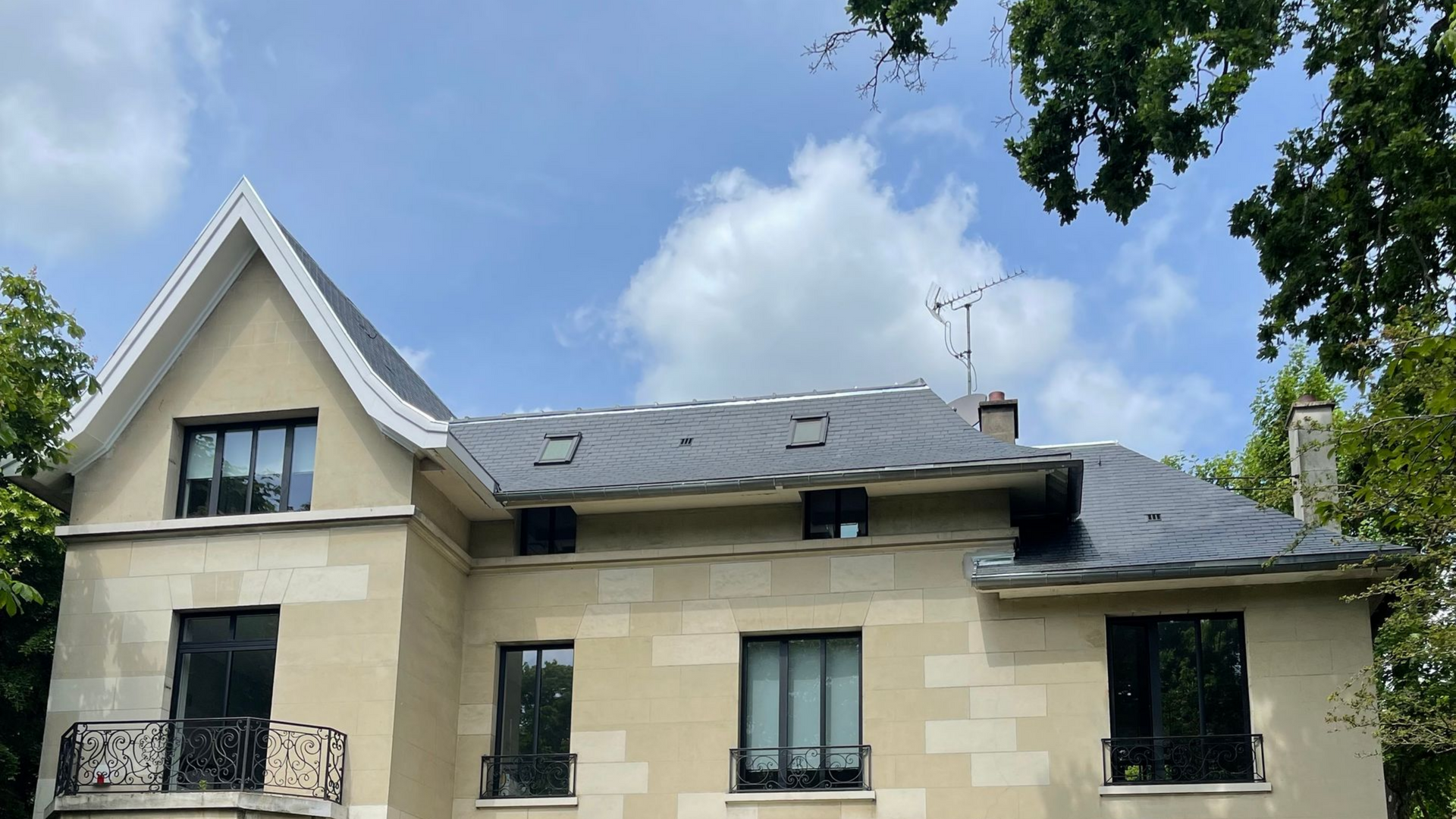 Maison en pierre beige au toit et aux fenêtres sombres, se détachant sur un ciel bleu nuageux. Des arbres encadrent la scène.