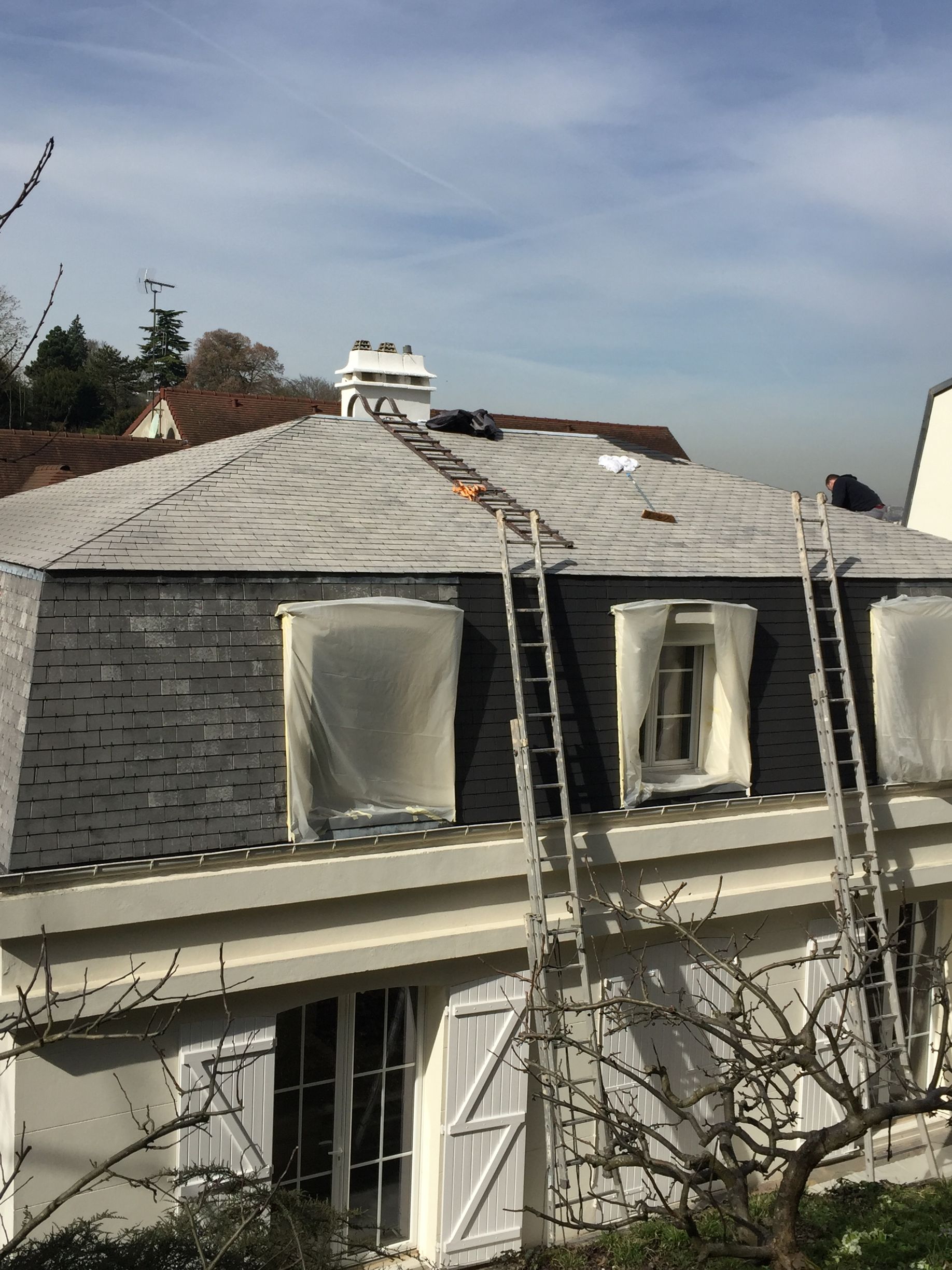 Deux échelles sont appuyées contre un toit d'ardoise avec une cheminée ; fenêtres recouvertes de plastique, ciel bleu.