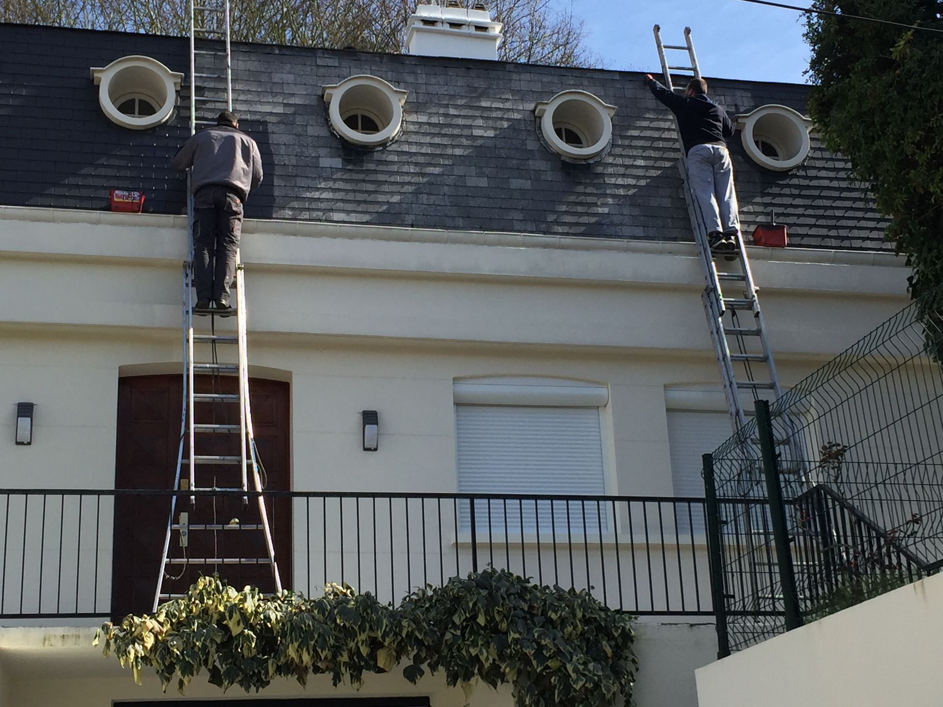 Deux ouvriers sur des échelles accèdent à un toit en ardoise percé de lucarnes circulaires, sur un bâtiment blanc.