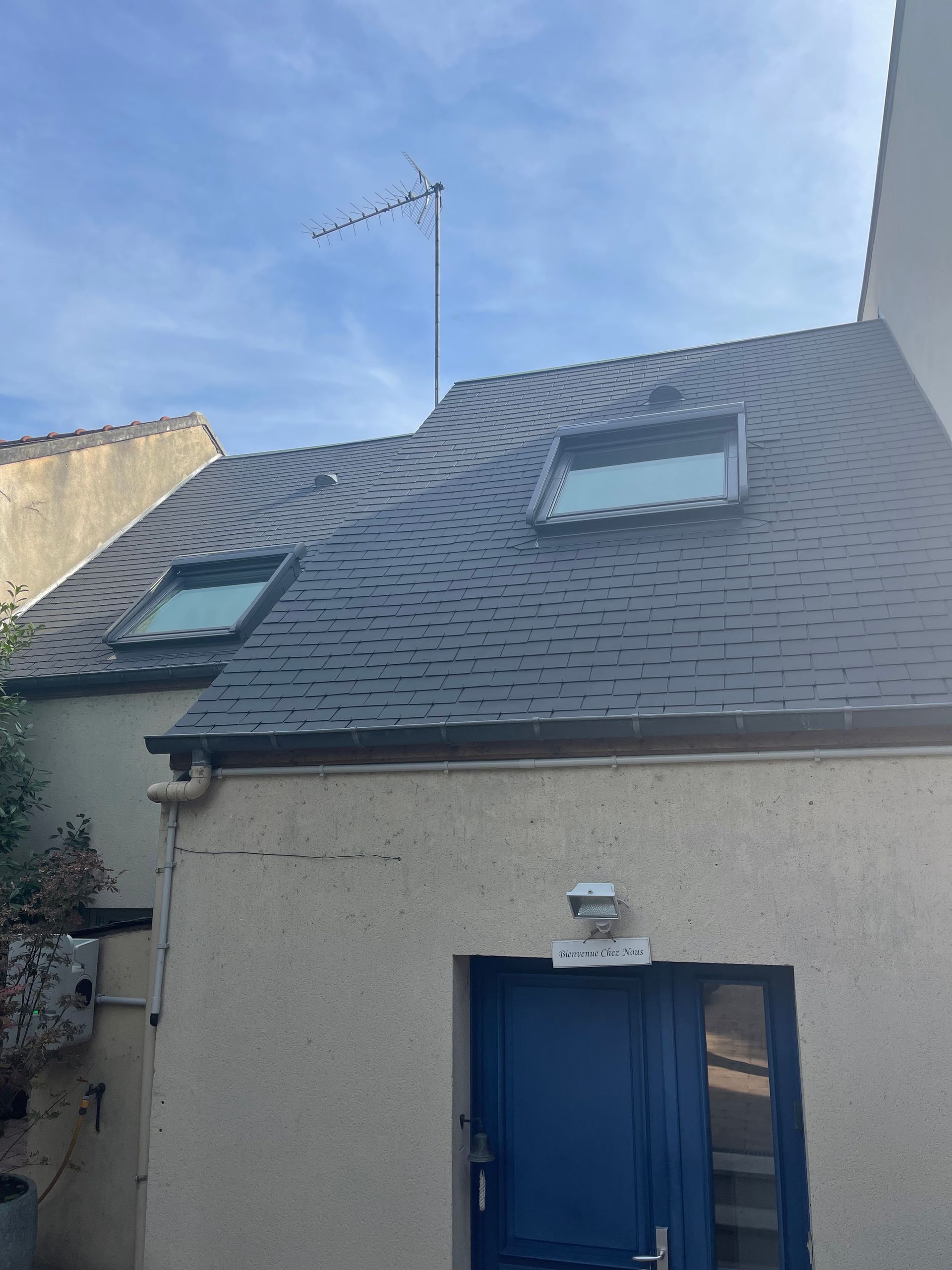 Bâtiment au toit sombre, percé de deux puits de lumière et doté d'une porte bleue. Une antenne de télévision est installée sur le toit.