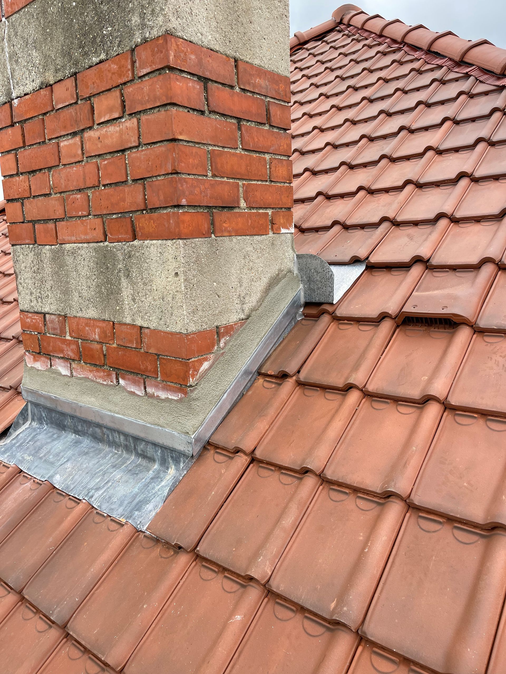 Cheminée en briques sur un toit de tuiles avec solins. Tuiles rouge-brun, ciment gris, ciel couvert.