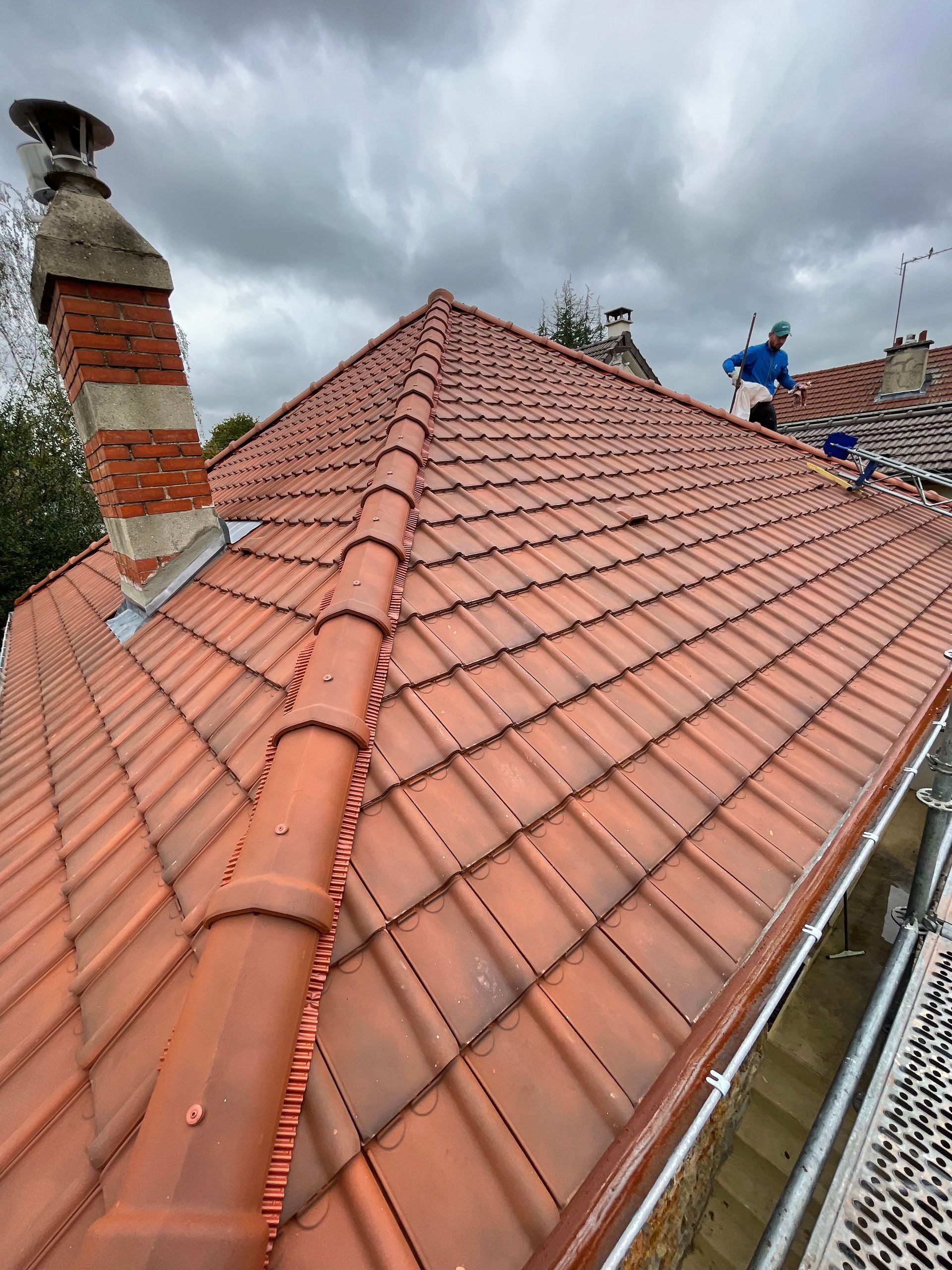 Toit de tuiles rouges avec cheminée en briques et gouttières se détachant sur un ciel nuageux.