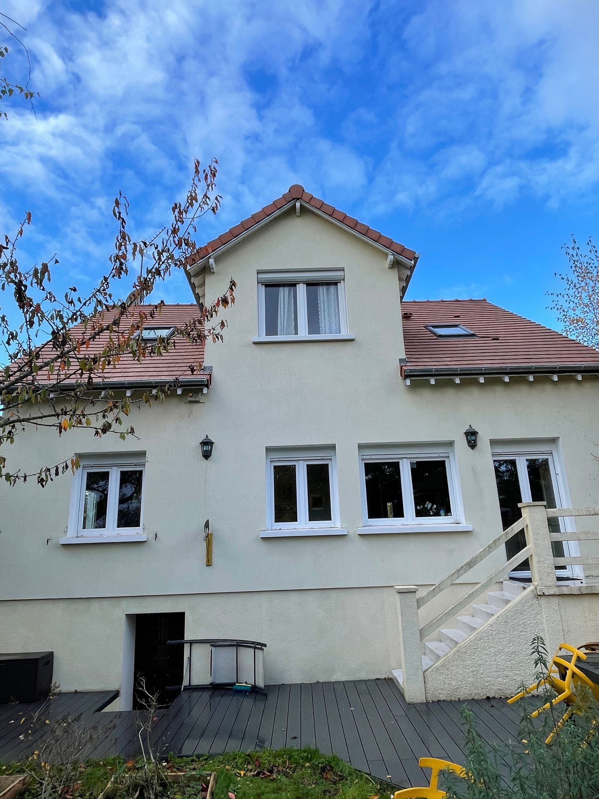 Maison beige au toit rouge, fenêtres blanches et ciel bleu. Un escalier mène à une terrasse latérale.