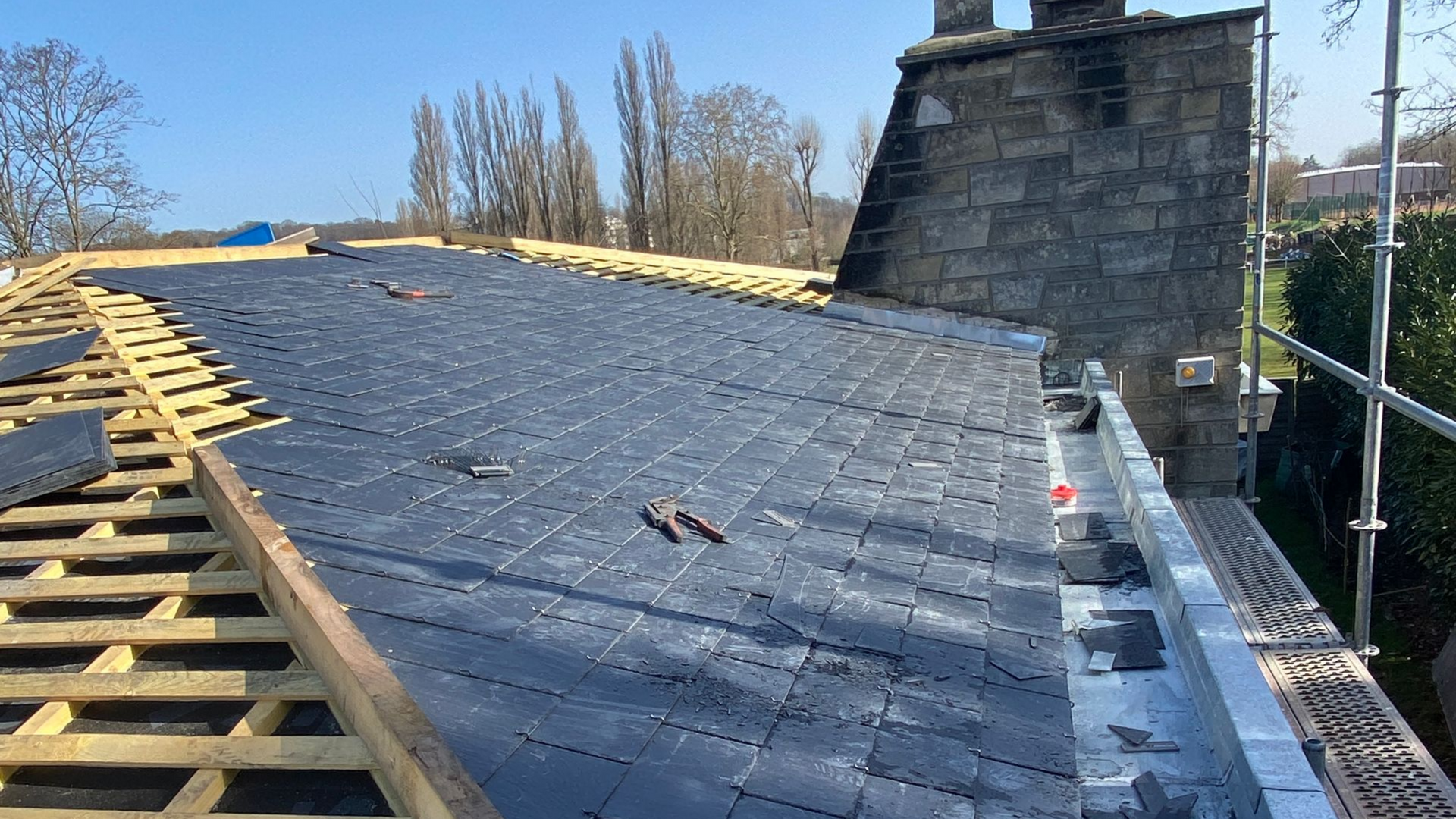 Toiture partiellement recouverte de tuiles gris foncé, poutres en bois apparentes. Échafaudages, cheminée et arbres en arrière-plan.