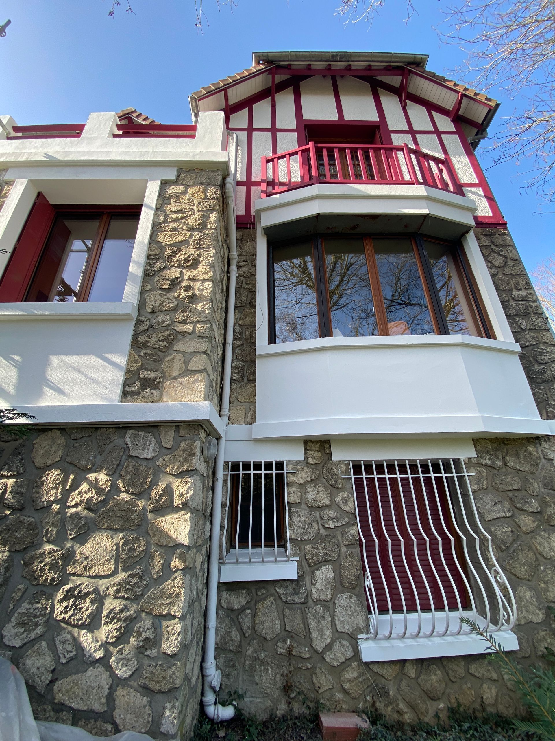 Maison en pierre de deux étages avec des boiseries rouges, un balcon et des touches de blanc, sous un ciel bleu.