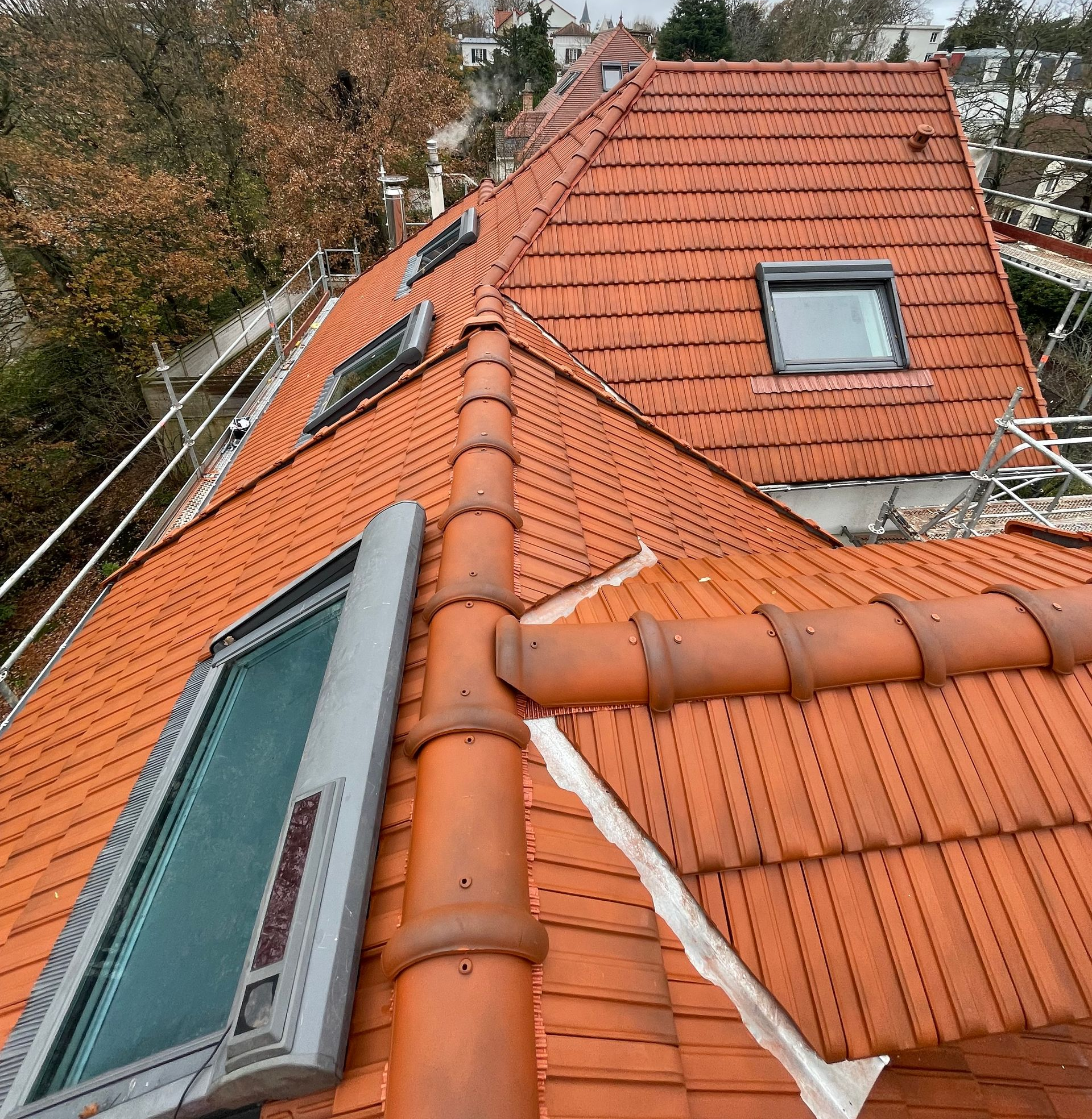 Toit de tuiles rouges percé de plusieurs puits de lumière, vu d'en haut, entouré d'arbres sous un ciel nuageux.