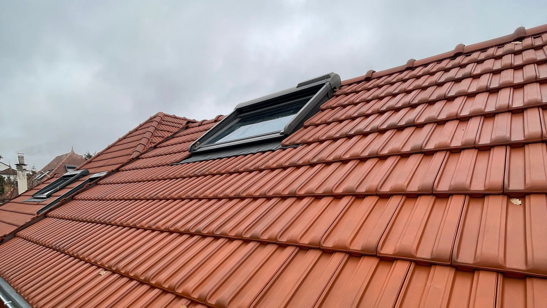 Toit de tuiles rouges avec deux puits de lumière sous un ciel nuageux.