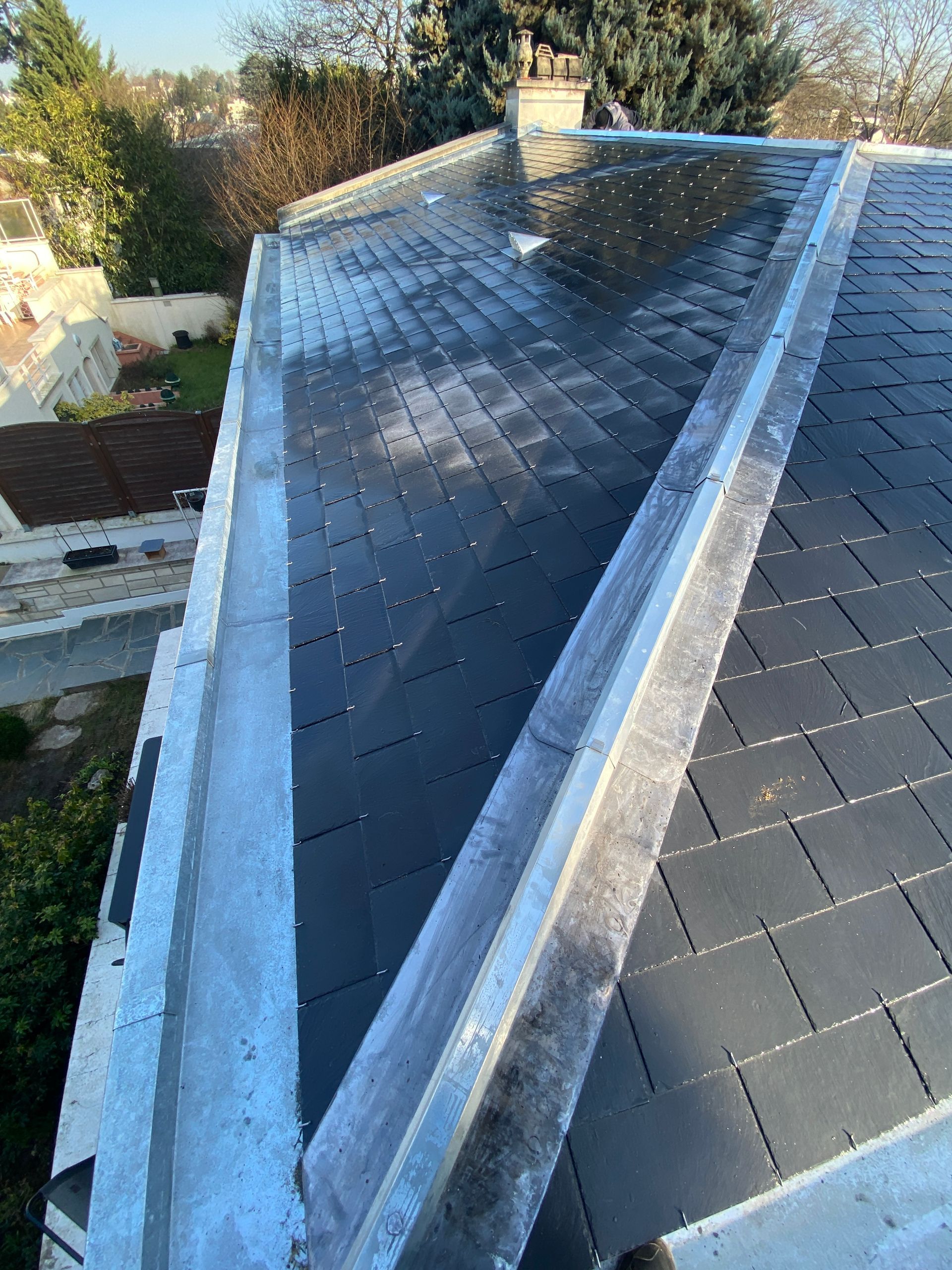 Vue aérienne d'un toit en ardoise avec une gouttière métallique. Le toit est partiellement ensoleillé. On aperçoit des arbres et des maisons.