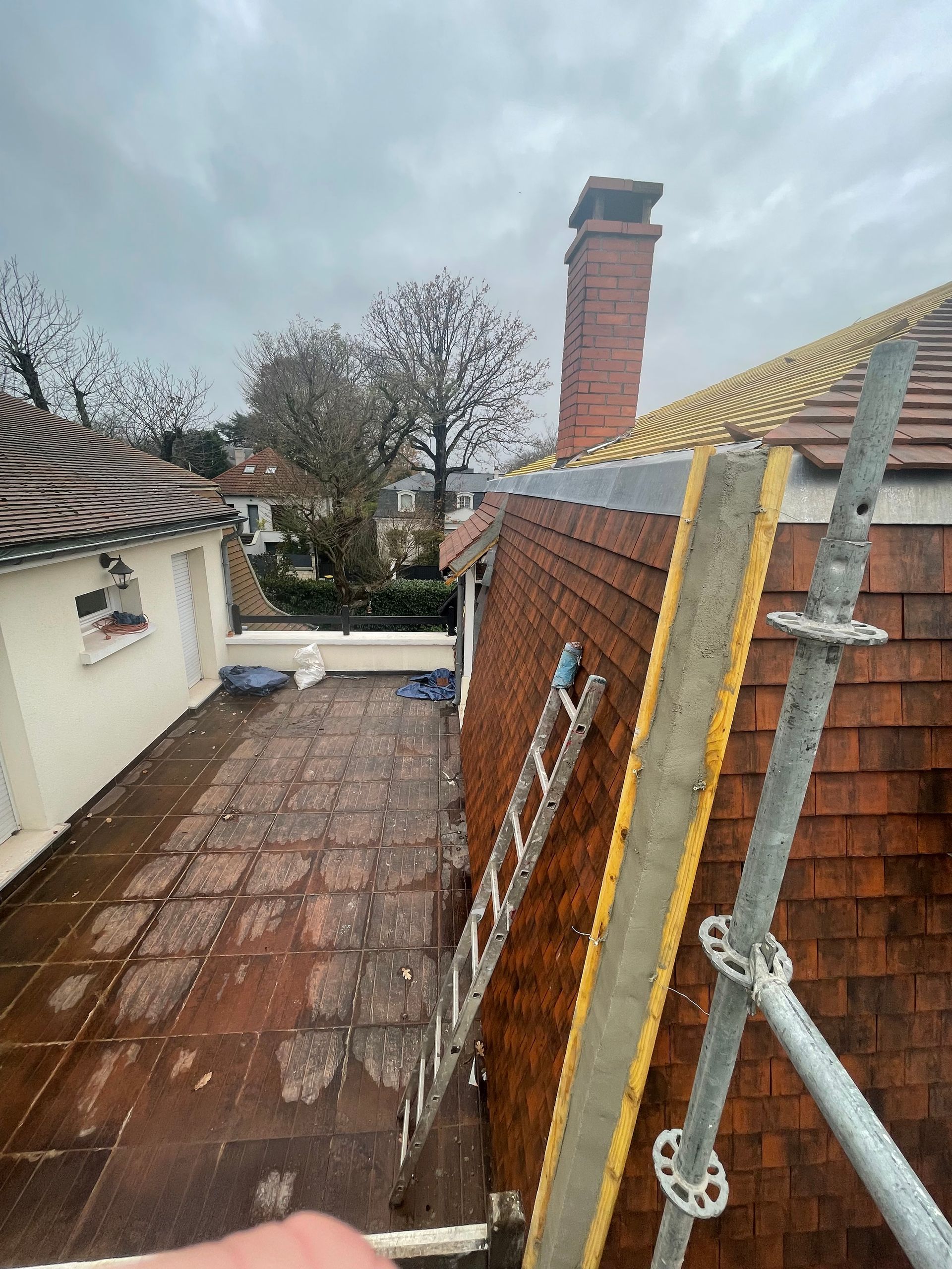 Vue du toit d'une maison avec un toit de tuiles rouges, une cheminée, une échelle et un échafaudage sur fond de ciel nuageux.