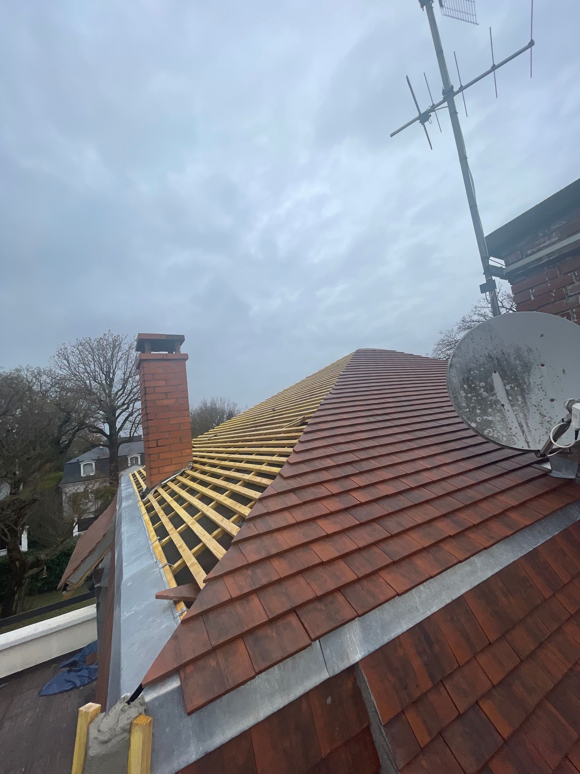 Toiture en construction, laissant apparaître des tuiles rouges, des poutres en bois, une cheminée, une antenne parabolique et un ciel couvert.