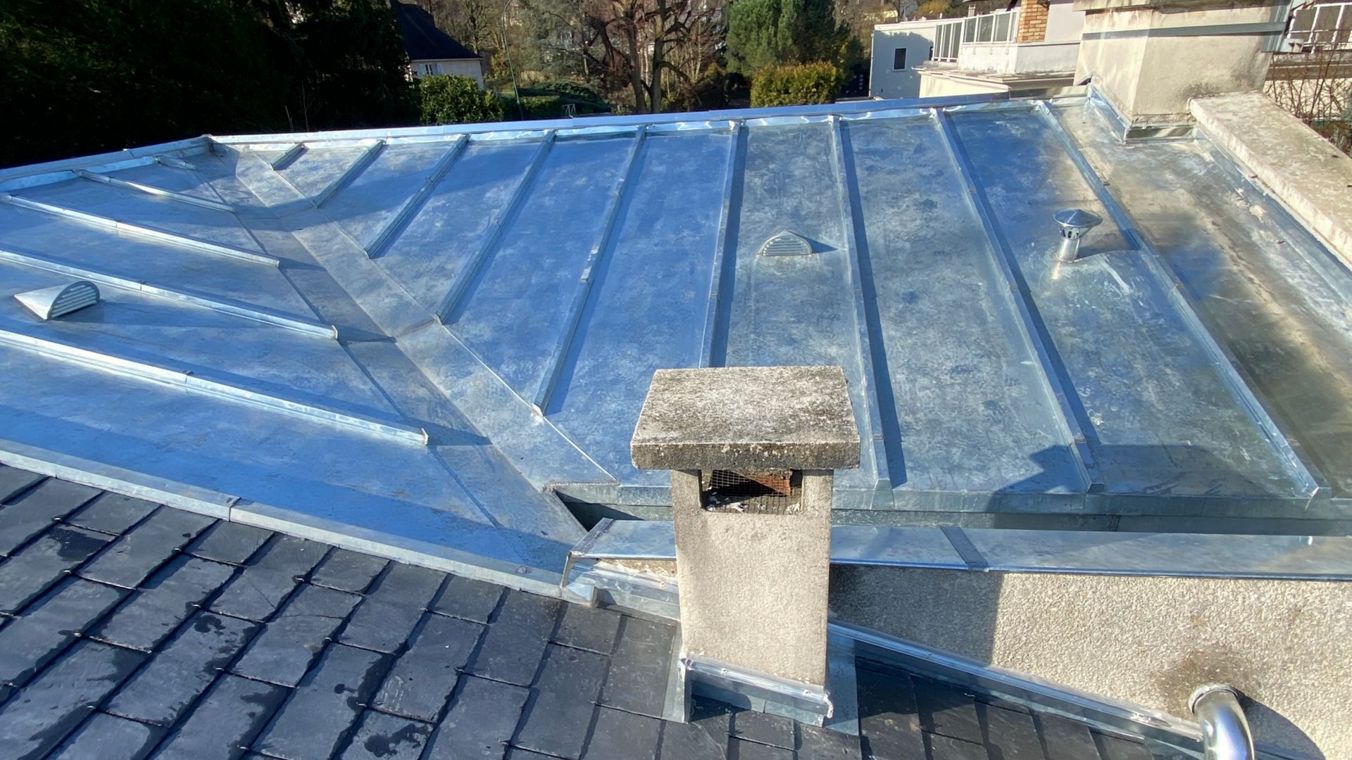 Gros plan sur le toit d'un bâtiment, avec sa toiture métallique brillante et sa cheminée. On aperçoit des tuiles sombres en dessous.