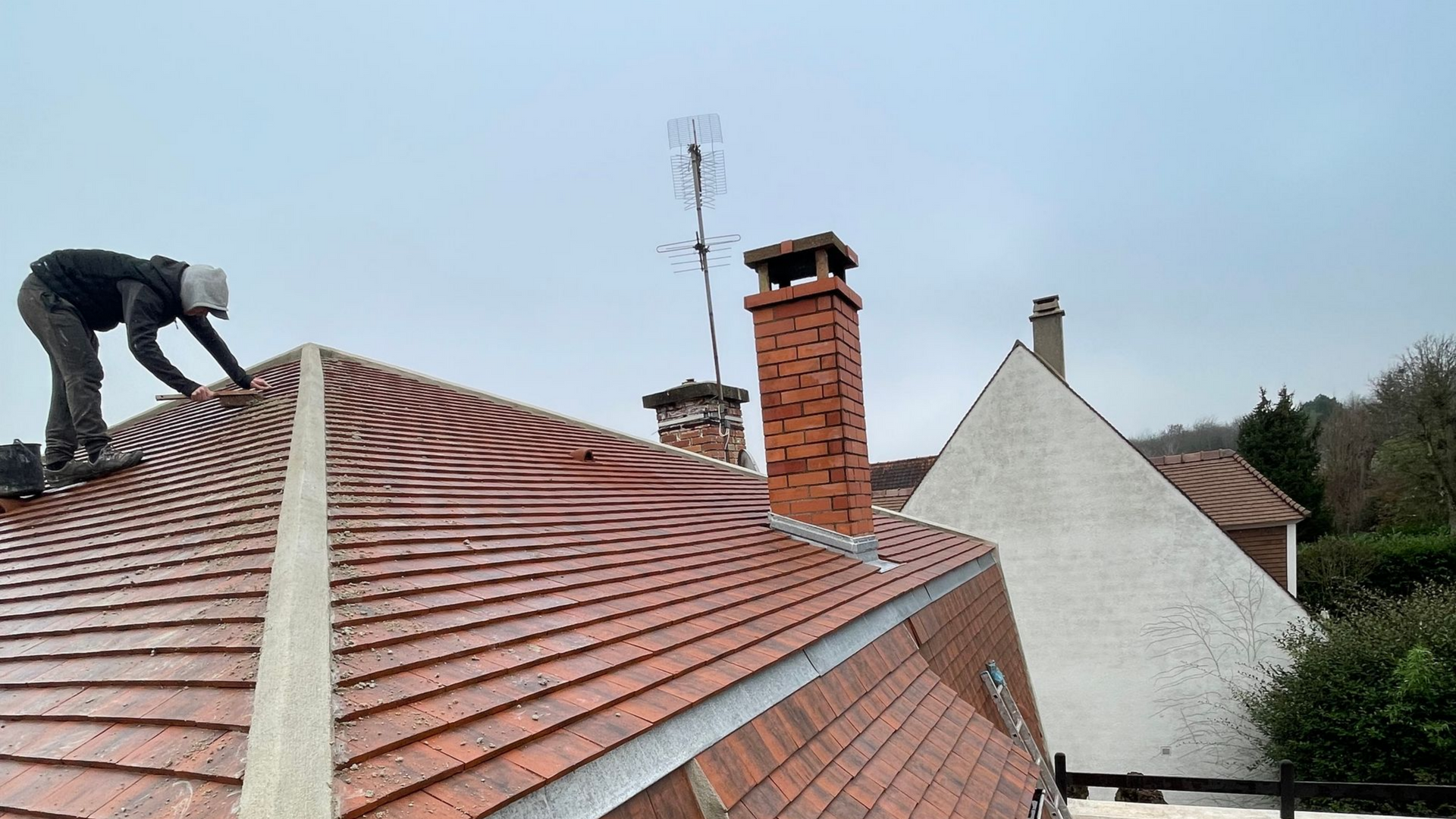 Une personne sur un toit de tuiles rouges, près d'une cheminée. Temps couvert.