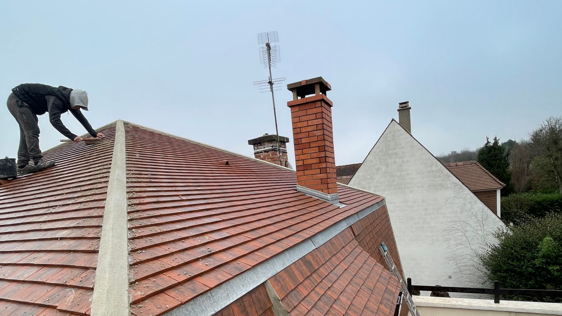 Couvreur sur un toit de tuiles en terre cuite, travaillant près d'une cheminée en briques et d'une antenne. Journée nuageuse.