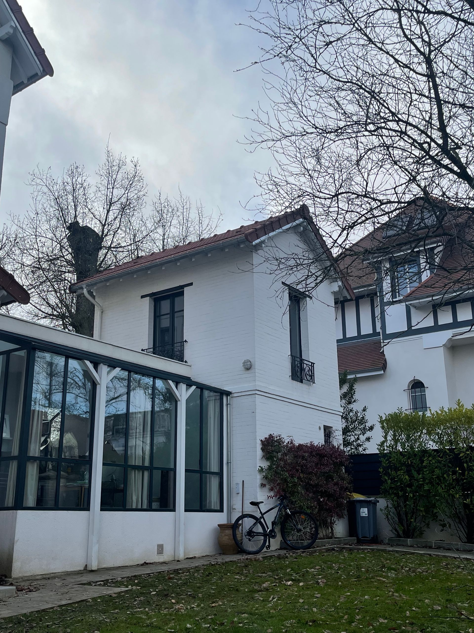 Bâtiment blanc au toit de tuiles rouges, extension aux parois de verre, vélo et arbres dénudés sur un ciel nuageux.