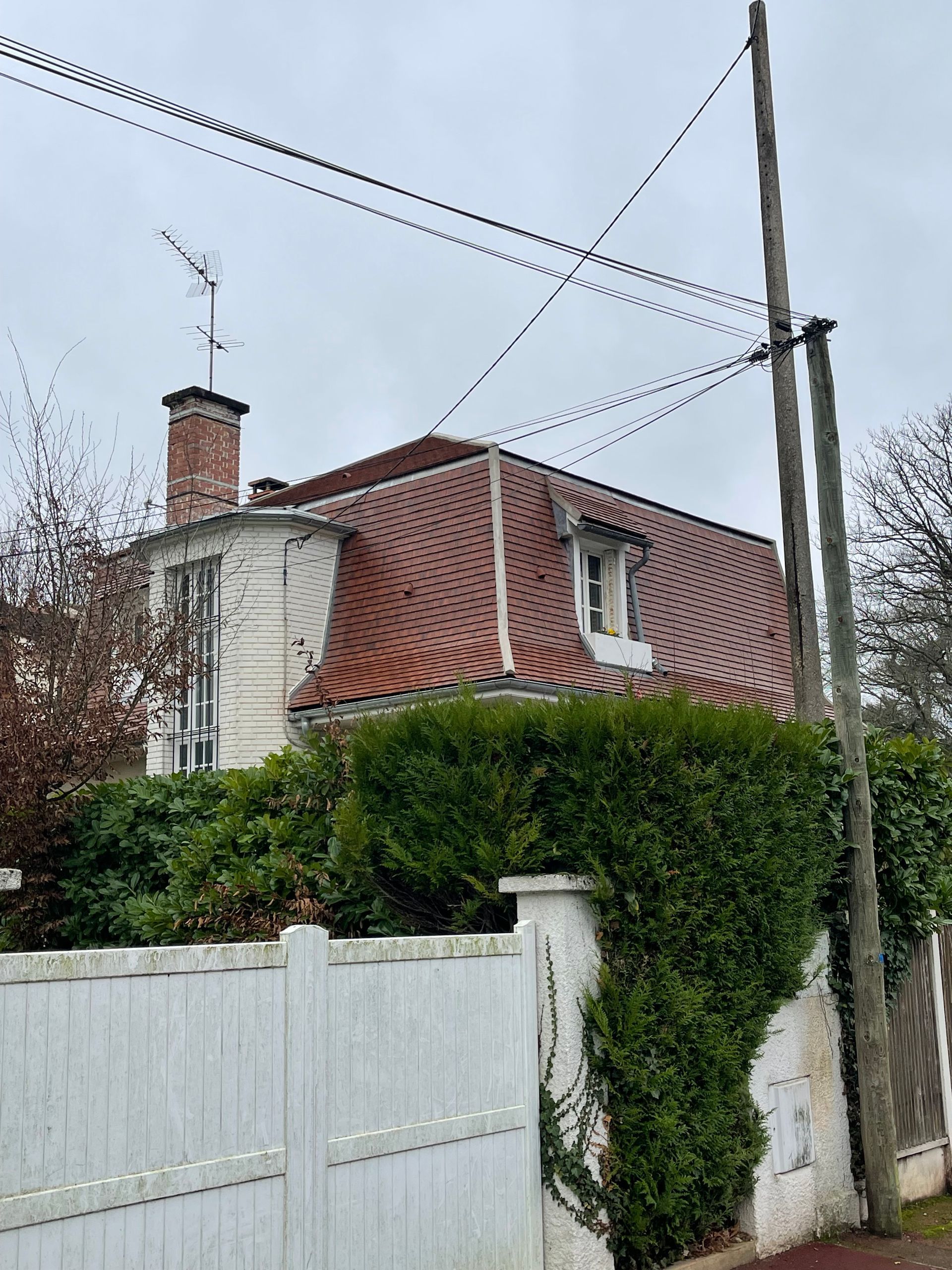 Maison blanche au toit de tuiles rouges, avec lucarne et cheminée. Buissons envahissants devant, ciel couvert.
