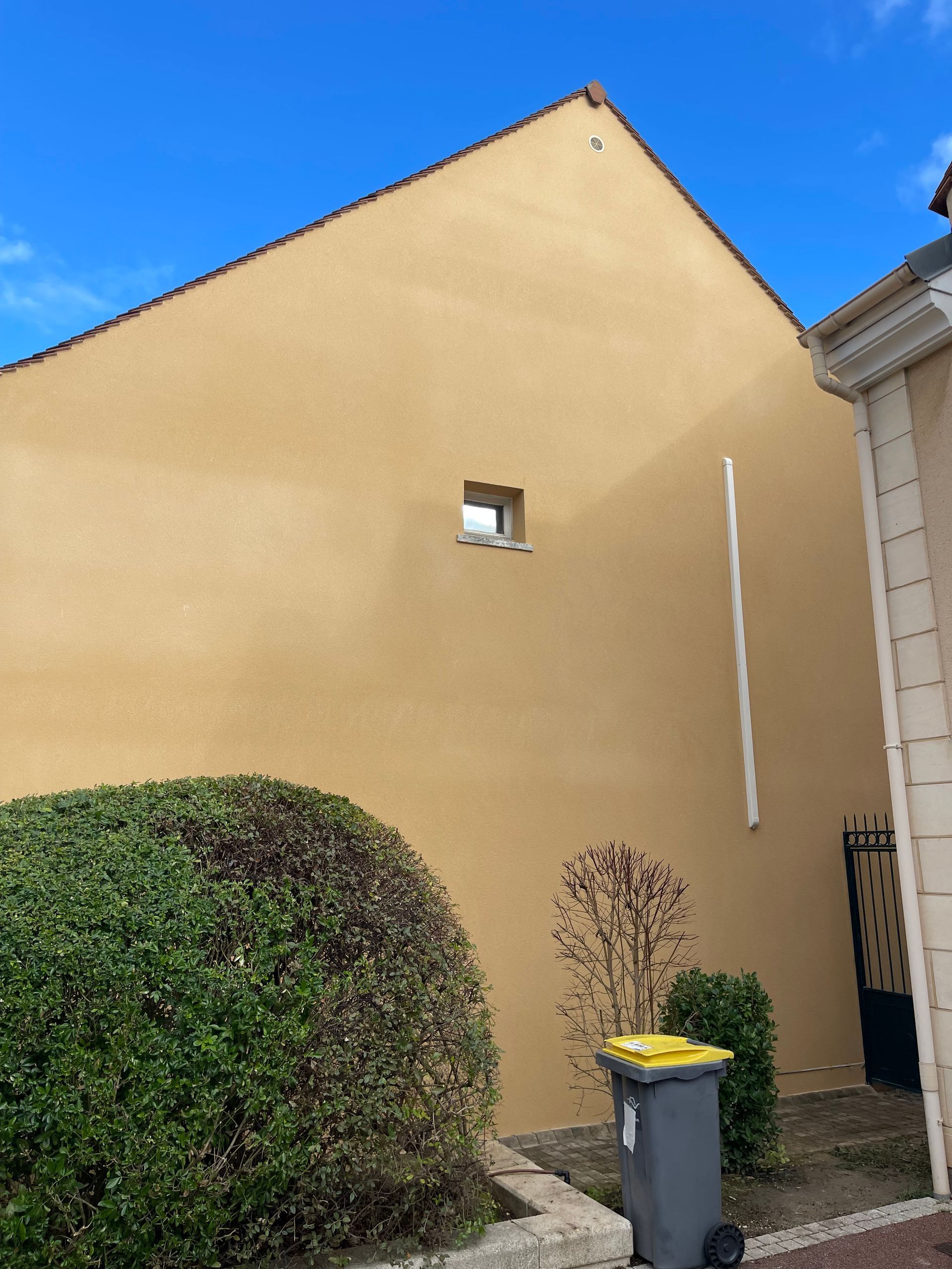 Maison jaune avec petite fenêtre, haie, poubelle et ciel bleu.
