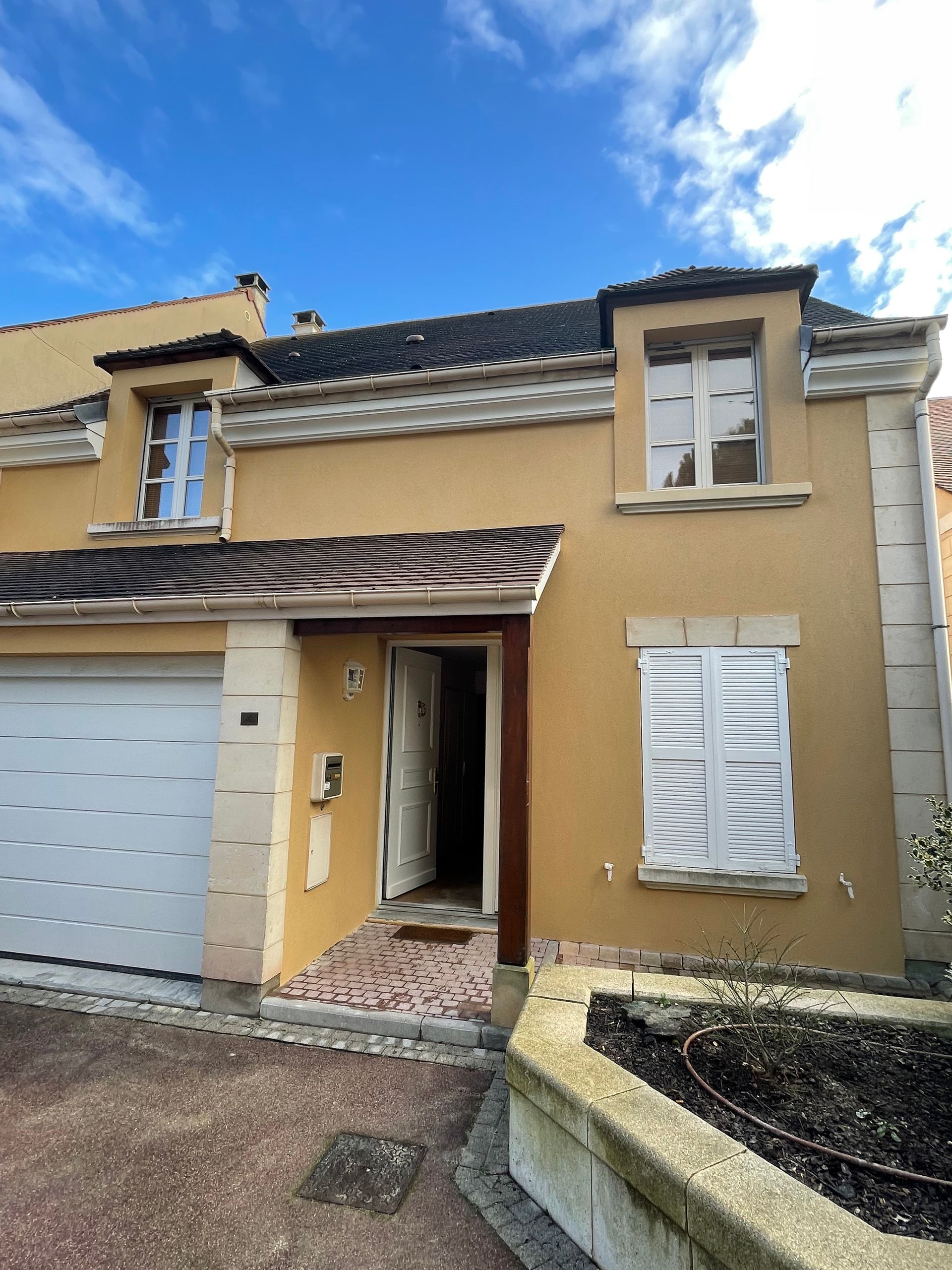 Maison beige à deux étages avec porte de garage et porte d'entrée ouverte ; volets blancs et ciel bleu.